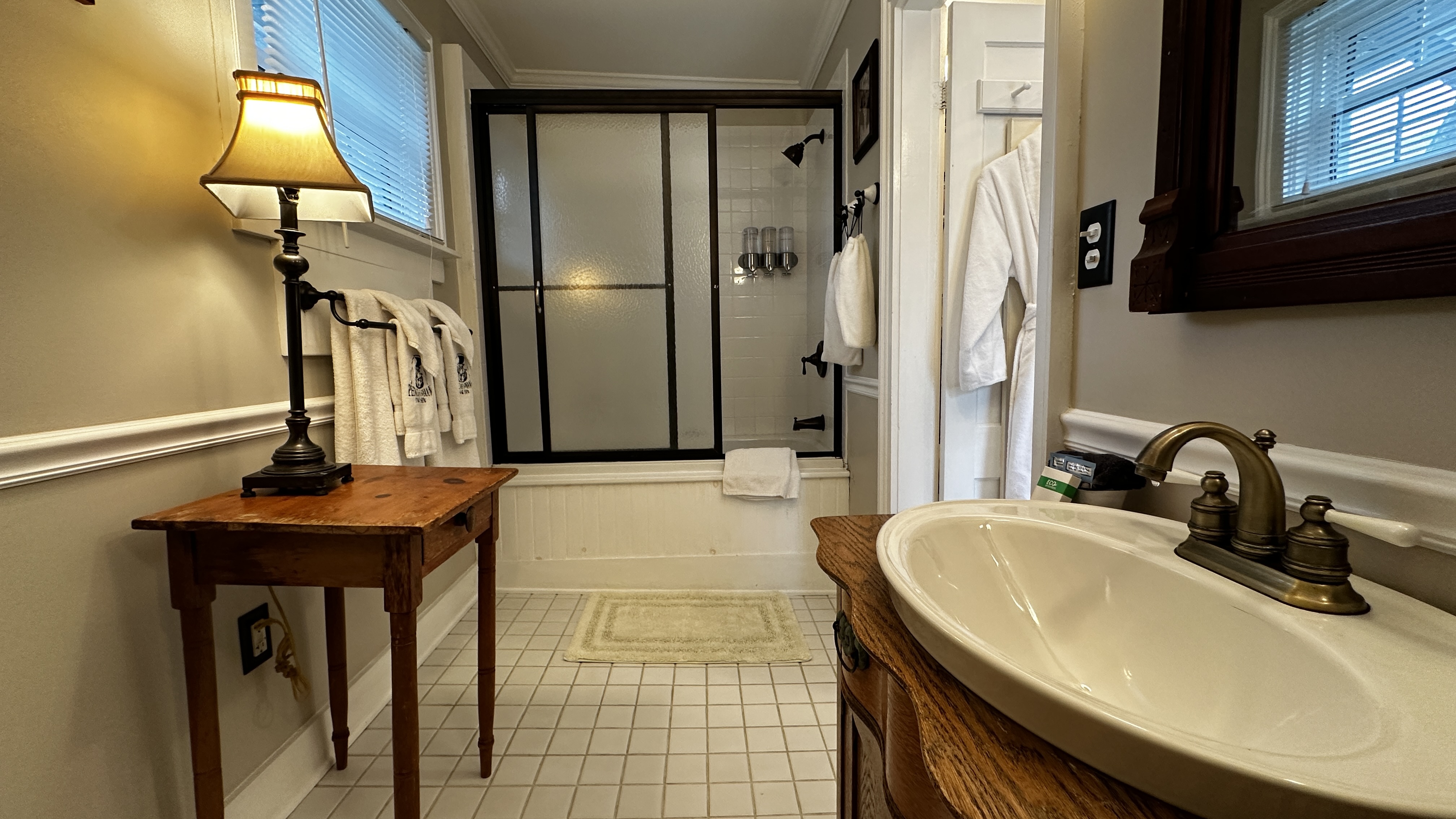 A classic bathroom featuring a white vessel sink on a rich wood vanity, an antique side table with a lamp, and a bathtub enclosed by black-framed frosted glass doors.