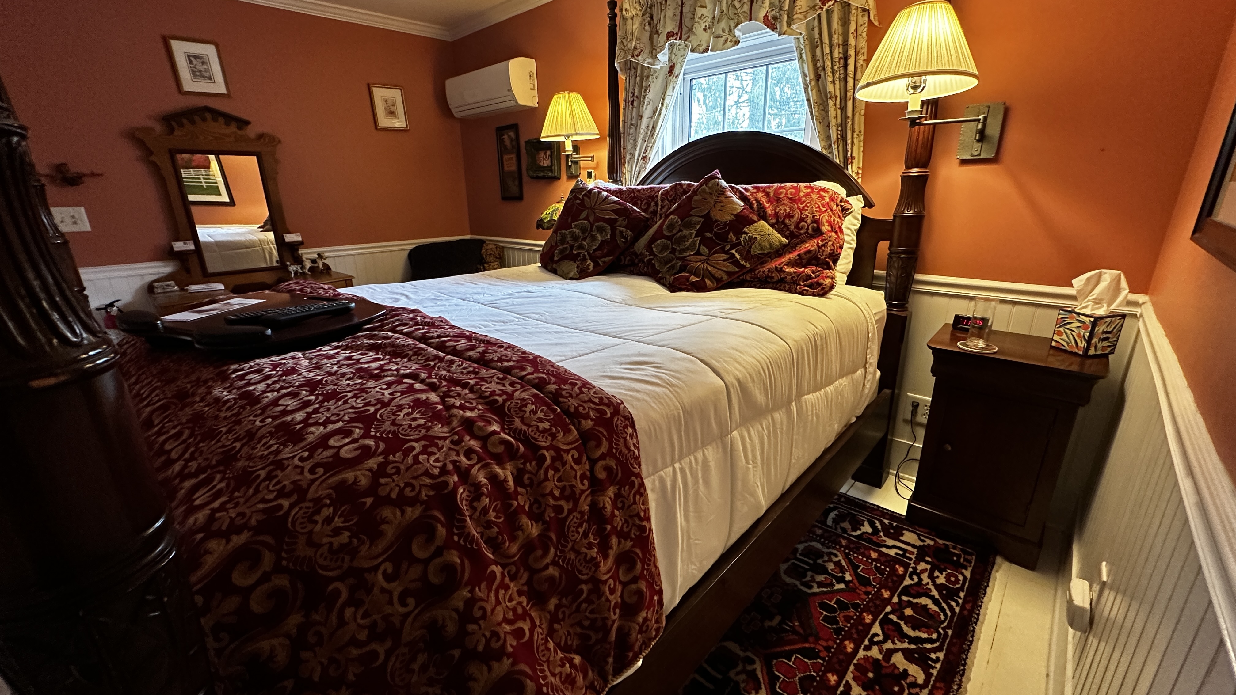 A cozy guest room with burnt sienna red walls and white wainscoting, featuring a dark wood queen-size four-poster bed with white linens and a red patterned throw. An antique dresser with a mirror and a bedside lamp add to the warm, traditional aesthetic.
