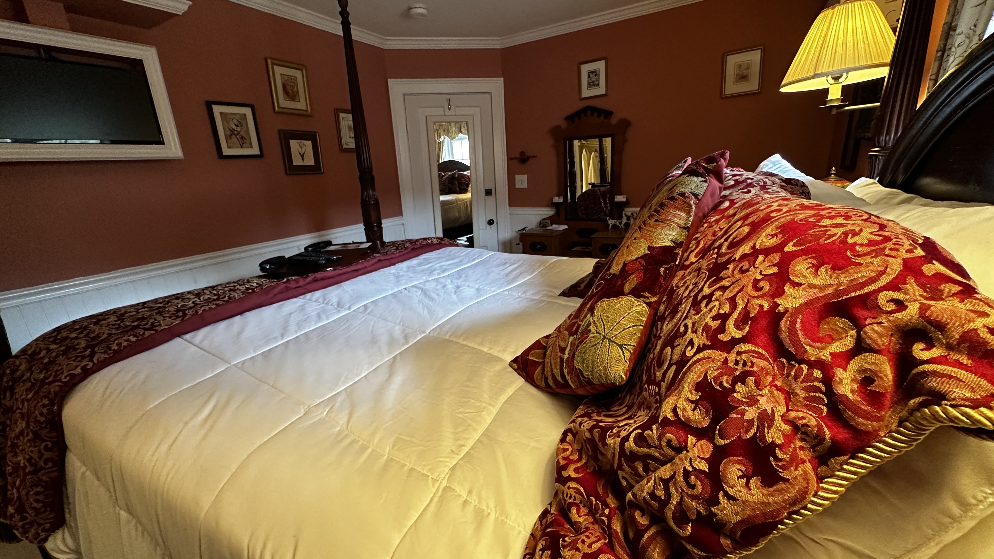 A guestroom with a dark wood four-poster bed with crisp white linens and a decorative red patterned throw. In the background, an antique wood dresser with an ornate mirror sits against burnt sienna walls with white wainscoting.