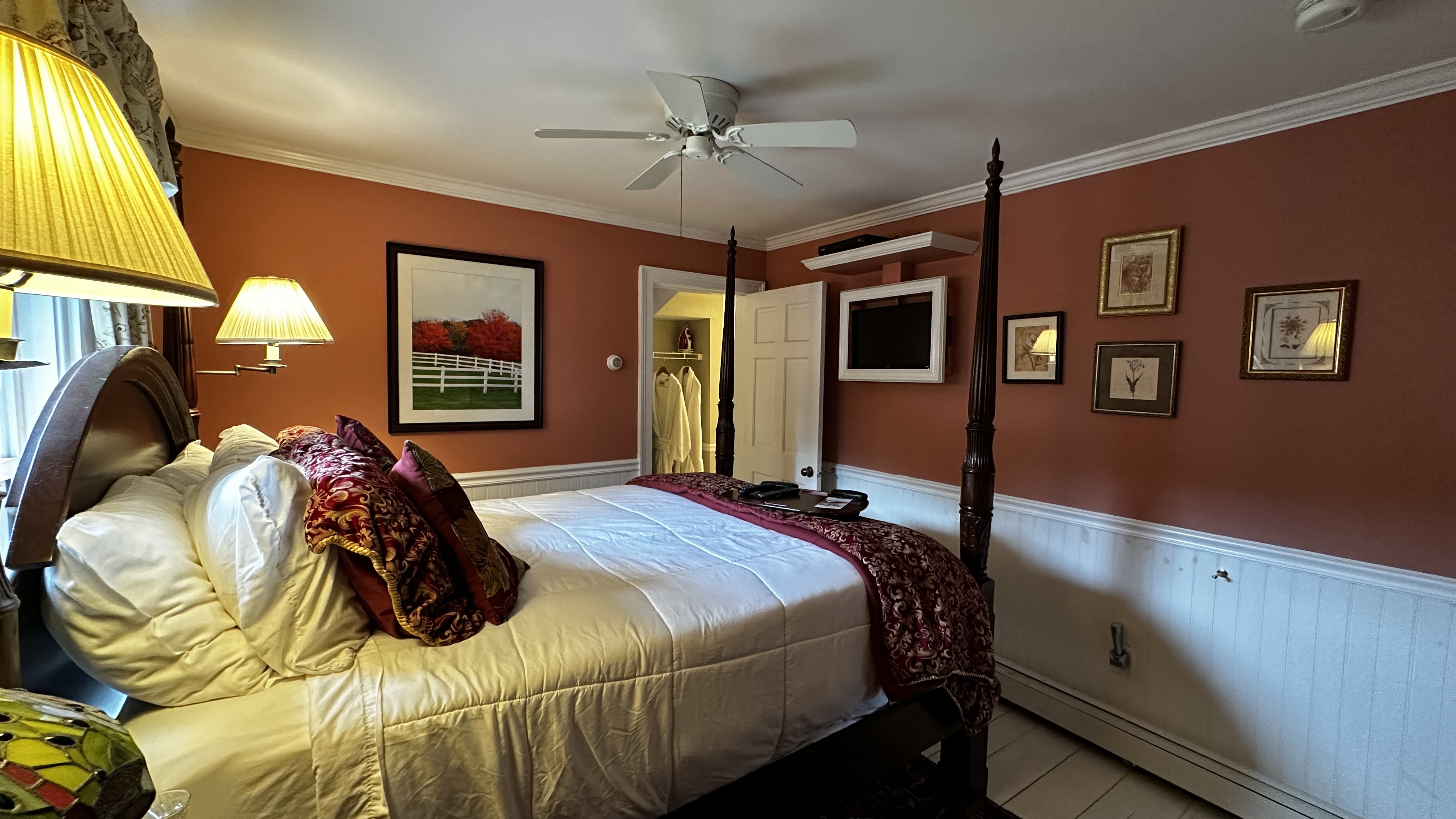 Red Room guest room with a dark wood queen-size four-poster bed with crisp white linens and a decorative red patterned throw. The room features burnt sienna walls above white beaded wainscoting, several framed pictures, and a wall-mounted flat-screen TV.