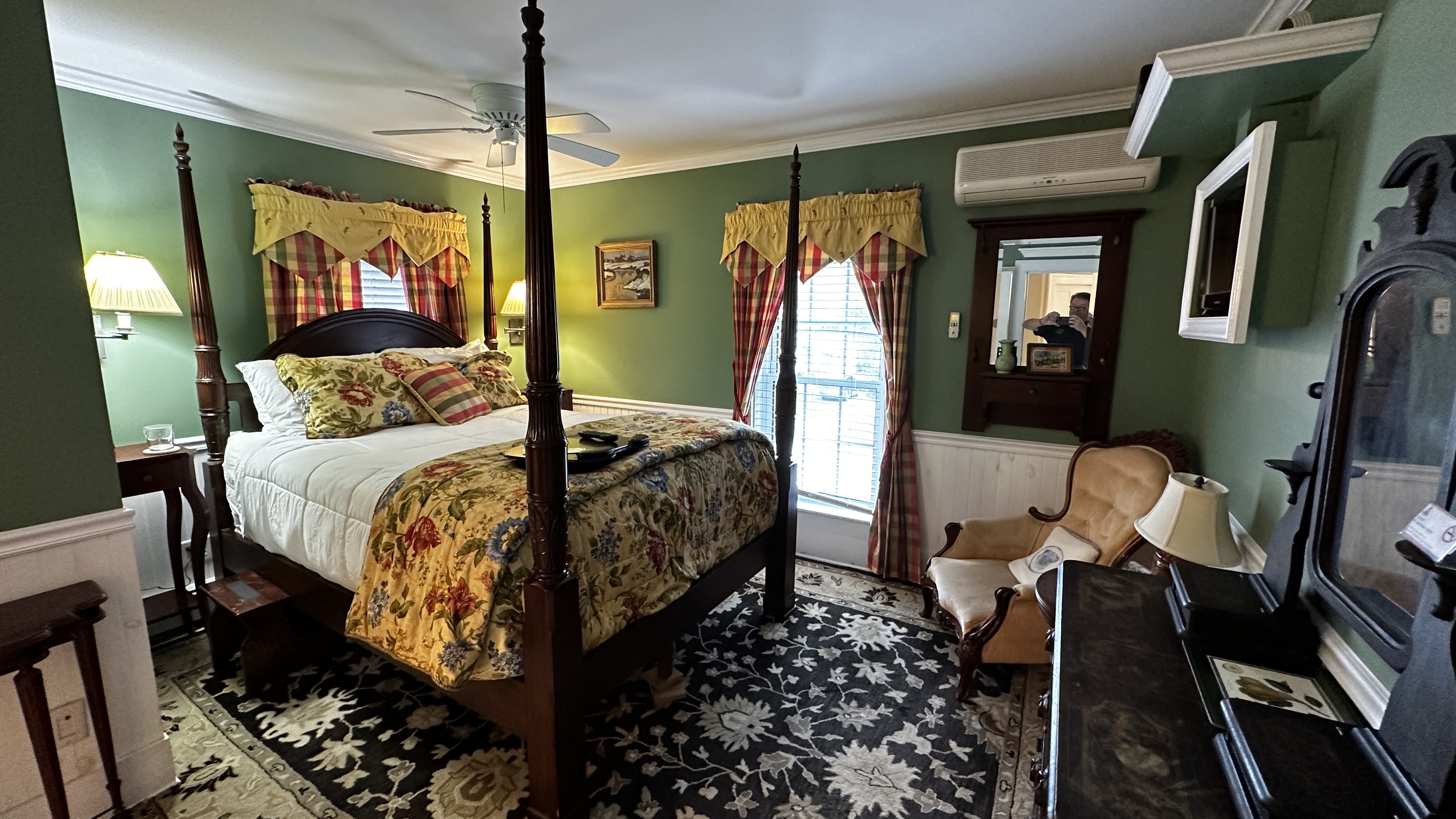 A traditional guest bedroom with fern-green walls, featuring a dark wood four-poster bed with a yellow floral coverlet. The room includes an antique dresser with an ornate mirror, a wingback chair, and a patterned area rug over wood floors.