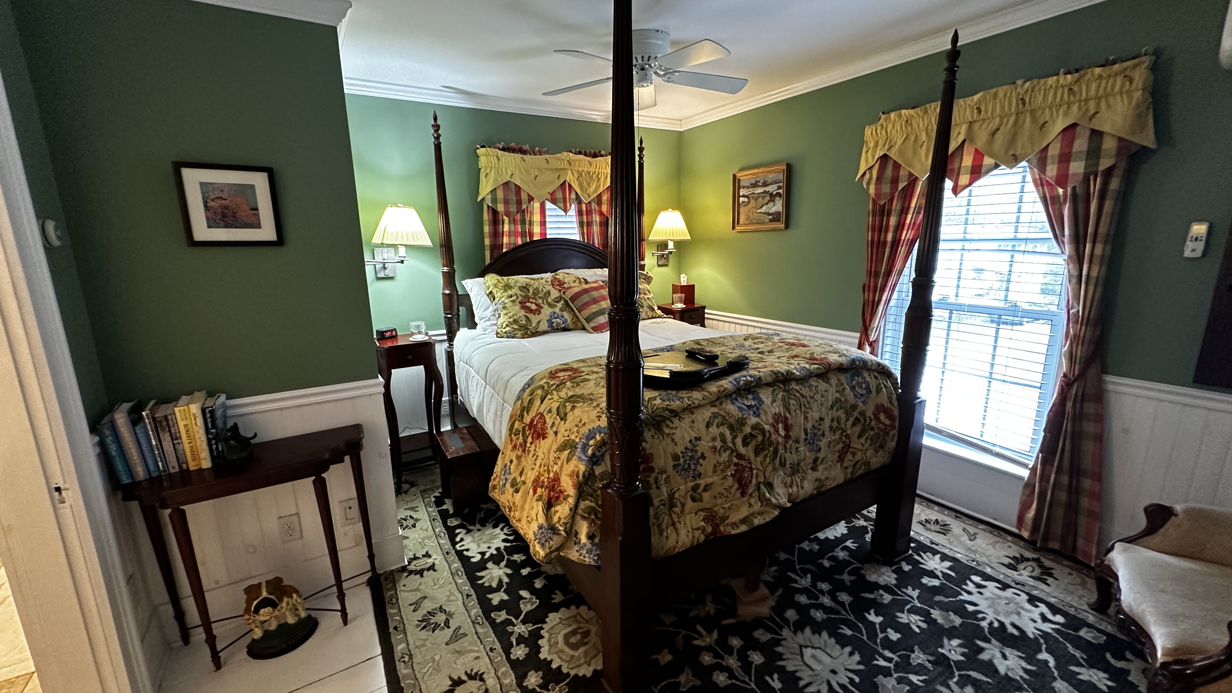 The traditional Green Room features fern-green walls, white wainscoting, and a dark wood four-poster bed with a yellow floral coverlet. The room is decorated with a patterned area rug, an antique dresser with an ornate mirror, and classic window valances.