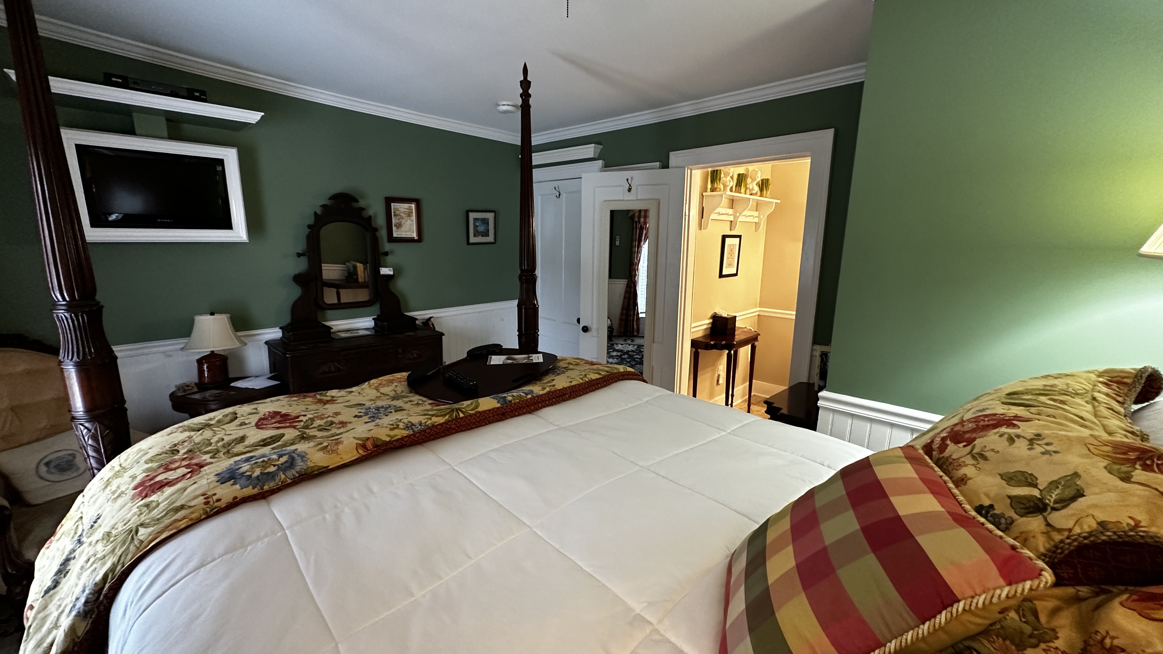 A guest room with fern-green walls, a dark wood four-poster bed with a yellow floral coverlet, and classic red-and-yellow plaid curtains. The room includes an antique dresser with an ornate mirror, a cream-colored armchair, and a dark patterned rug over white floors.
