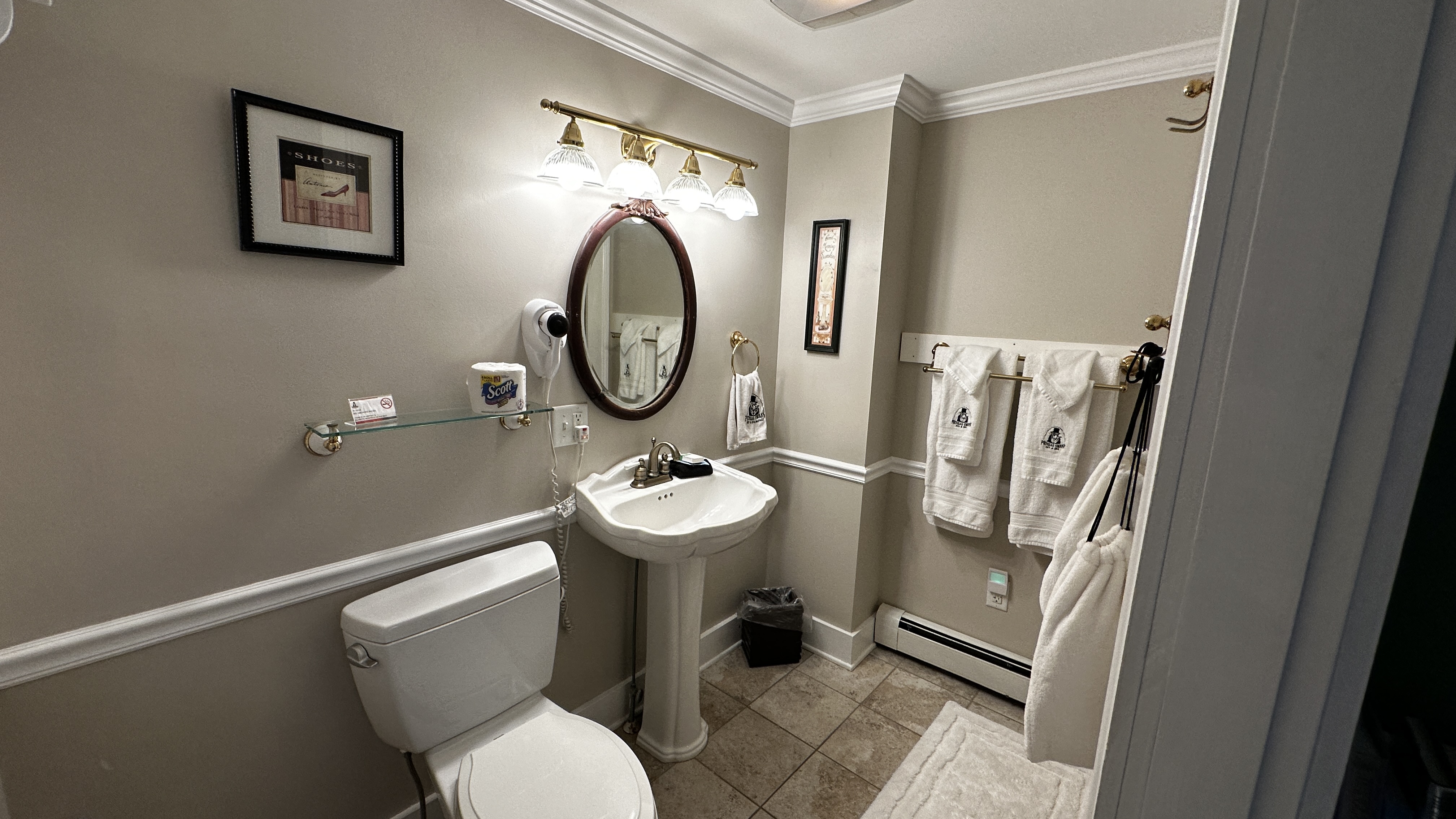 A traditional bathroom featuring a white pedestal sink with an oval mirror, a white toilet, and tan floor tiles. The walls are painted a neutral taupe with white wainscoting, and several white towels hang on a rack to the right.