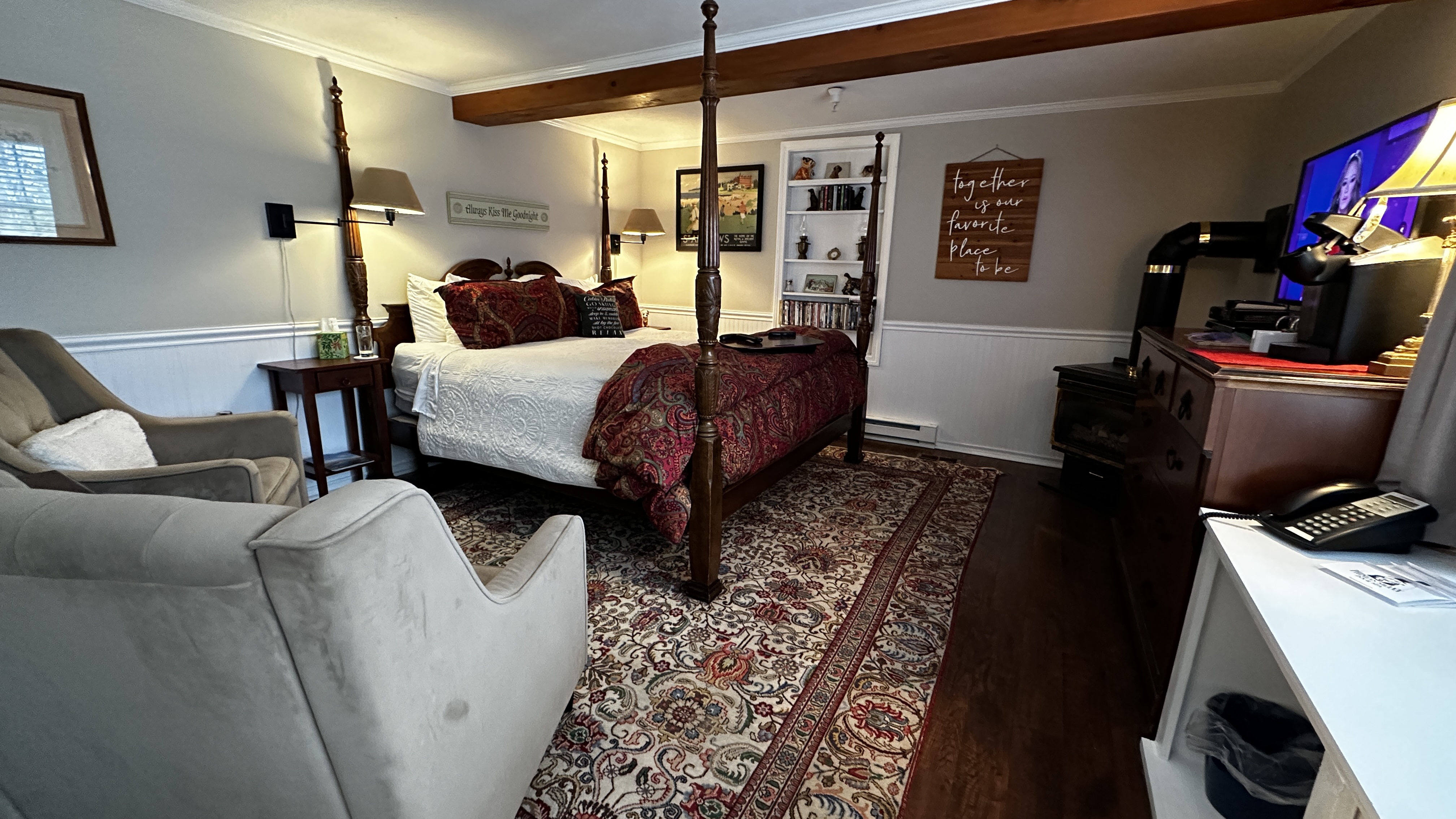 A spacious bedroom features a four-poster king-size bed with white linens and a red patterned throw. The room includes two plush gray reading chairs, a traditional patterned area rug over wood floors, and a warm, inviting atmosphere with a wall-mounted TV and decorative wall art.