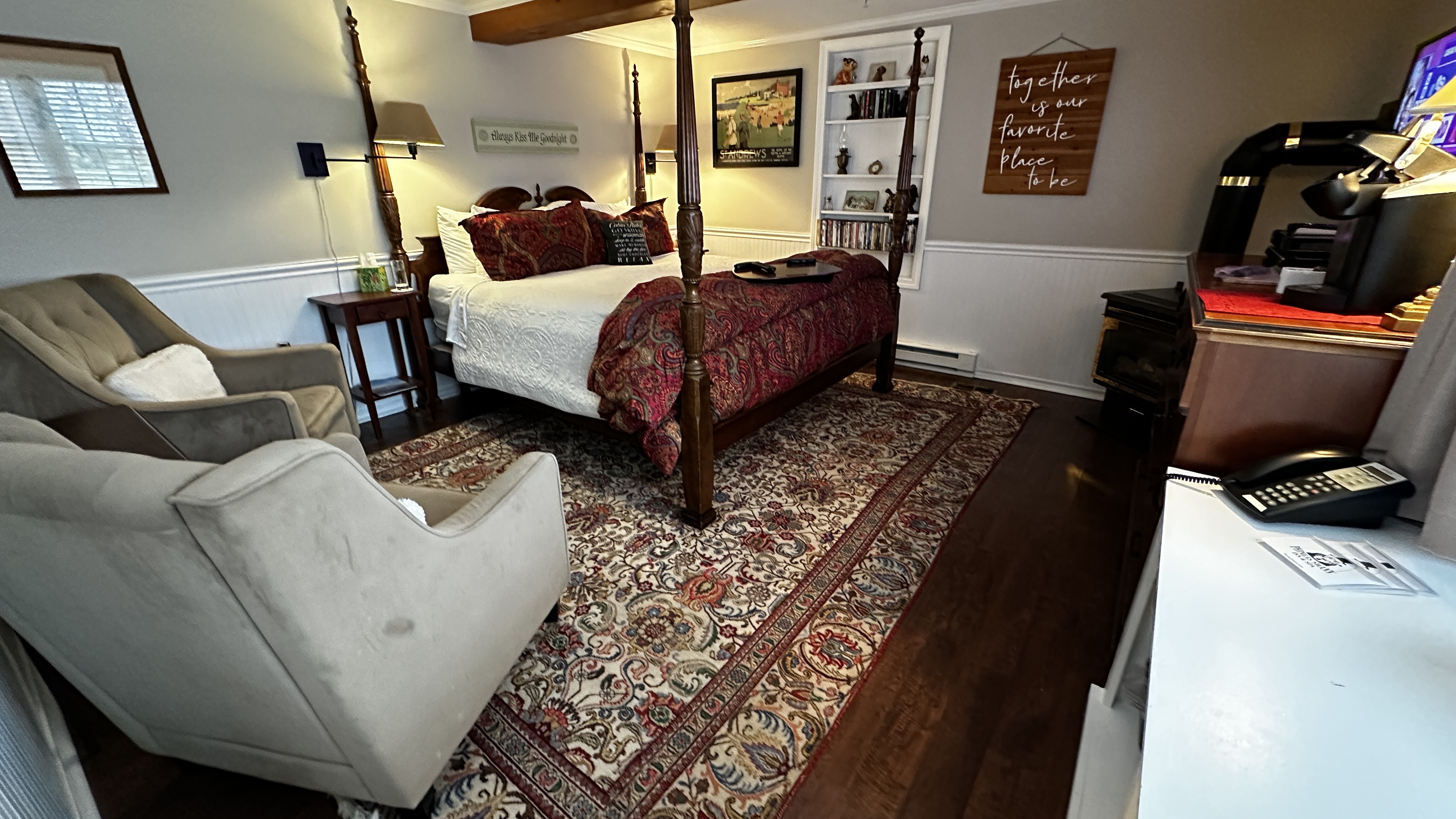 A spacious bedroom features a four-poster king-size bed with white linens and a red patterned throw. The room includes two plush gray reading chairs, a traditional patterned area rug over wood floors, and a warm, inviting atmosphere with a wall-mounted TV and decorative wall art.