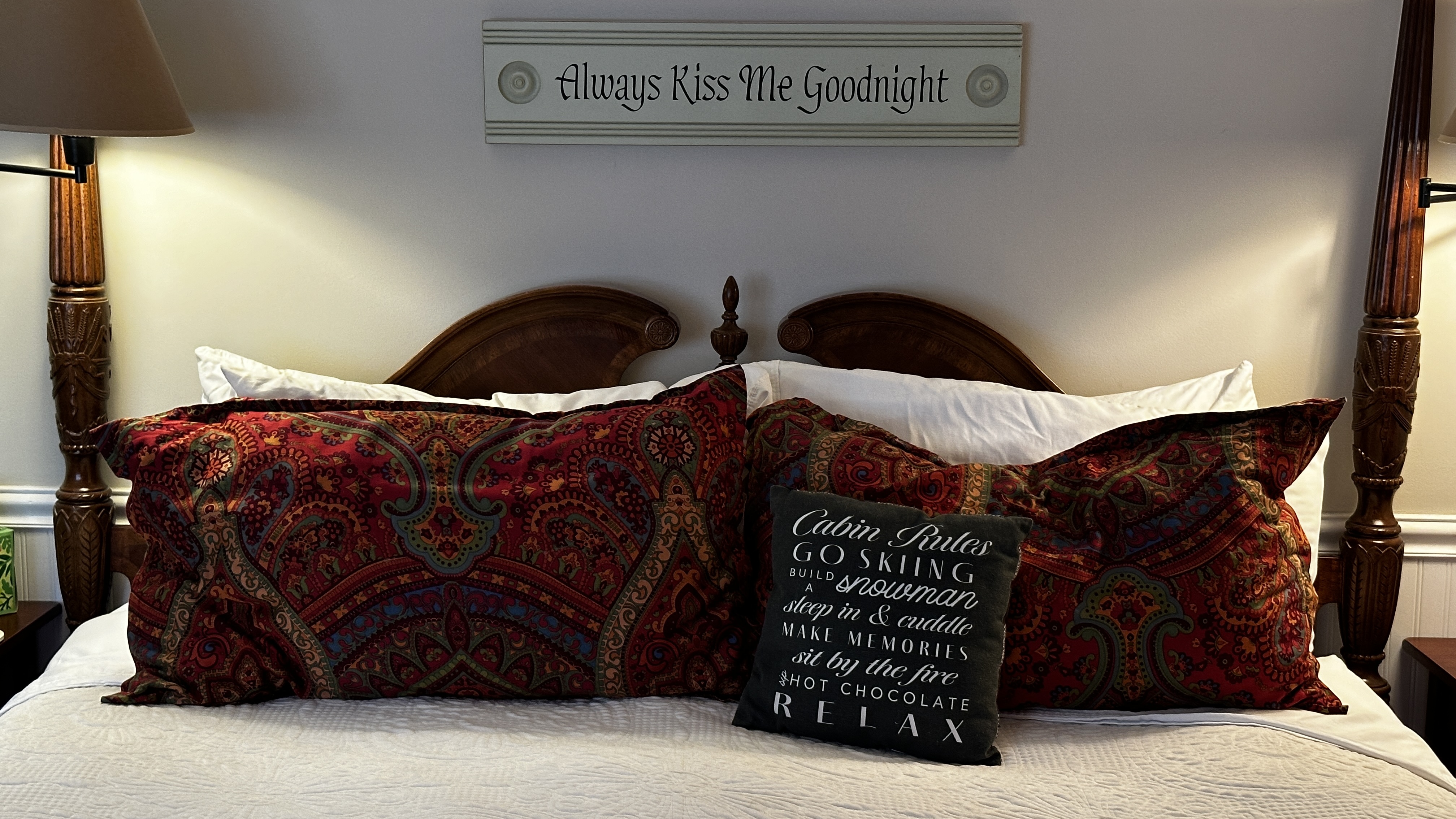 A close-up shot of a dark wood four-poster bed headboard with white linens and red patterned pillows. A small black decorative pillow in front features white text, and a sign above the bed reads, "Always Kiss Me Goodnight."