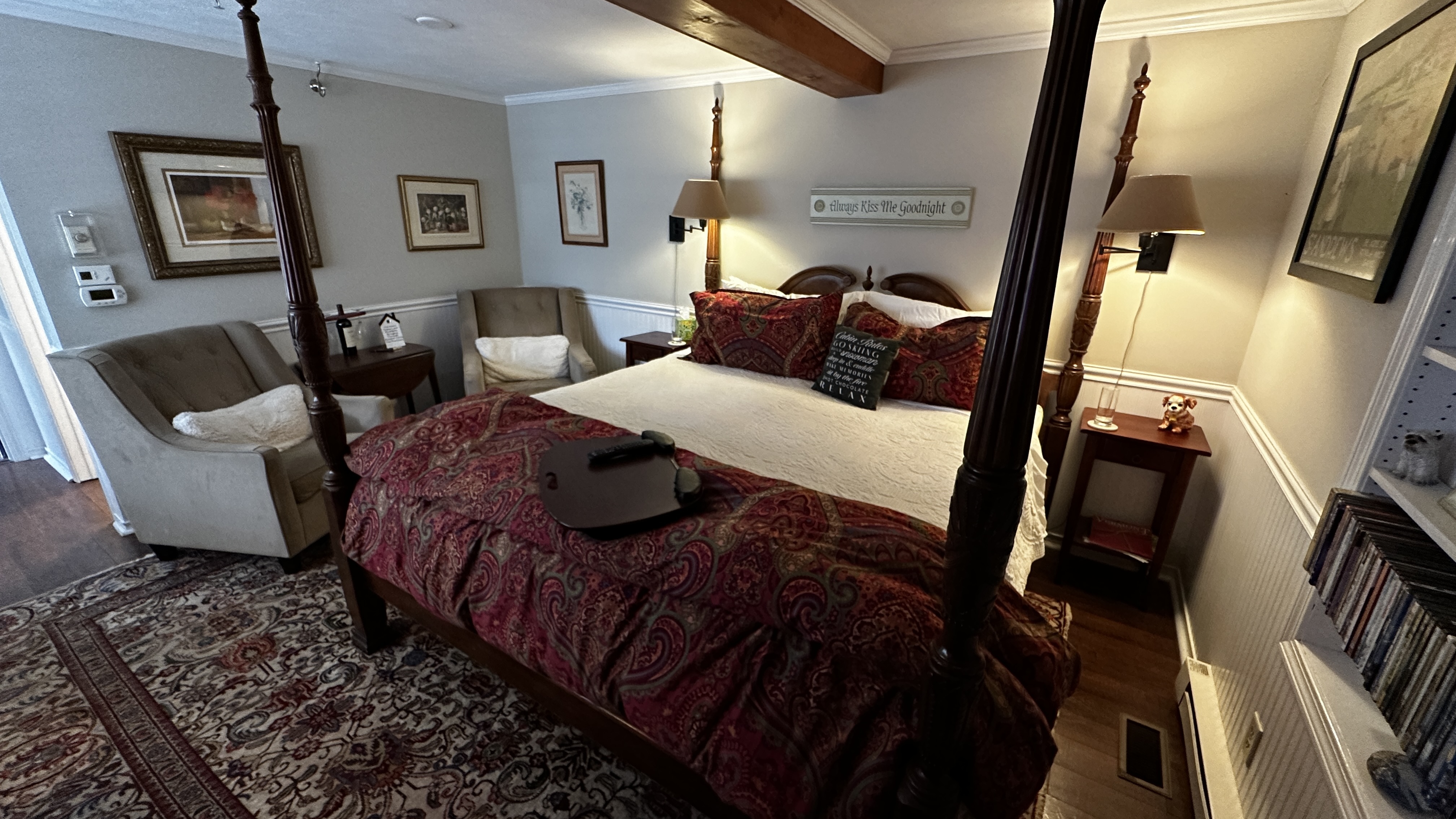 A cozy guest bedroom featuring a dark wood four-poster king-size bed with crisp white linens and a decorative red paisley throw. The room includes two plush gray armchairs, a traditional patterned area rug over dark wood floors, and warm lighting from bedside lamps.