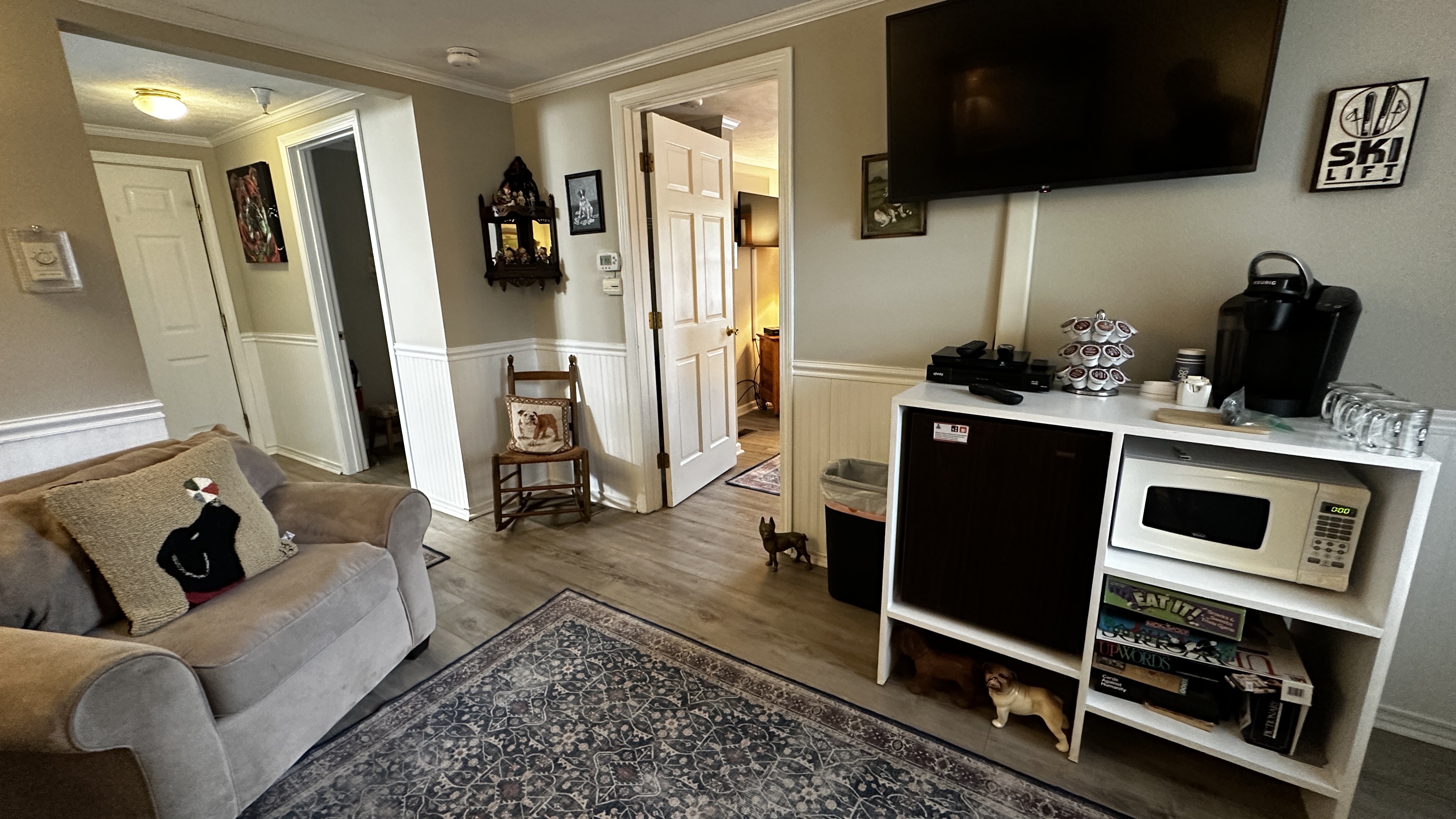 A living area featuring a light grey armchair with a black bear decorative pillow. The space includes a white storage unit equipped with a microwave, mini-fridge, and coffee maker, situated beneath a wall-mounted TV. In the background, doorways lead to other rooms and a hallway with a wooden rocking chair.