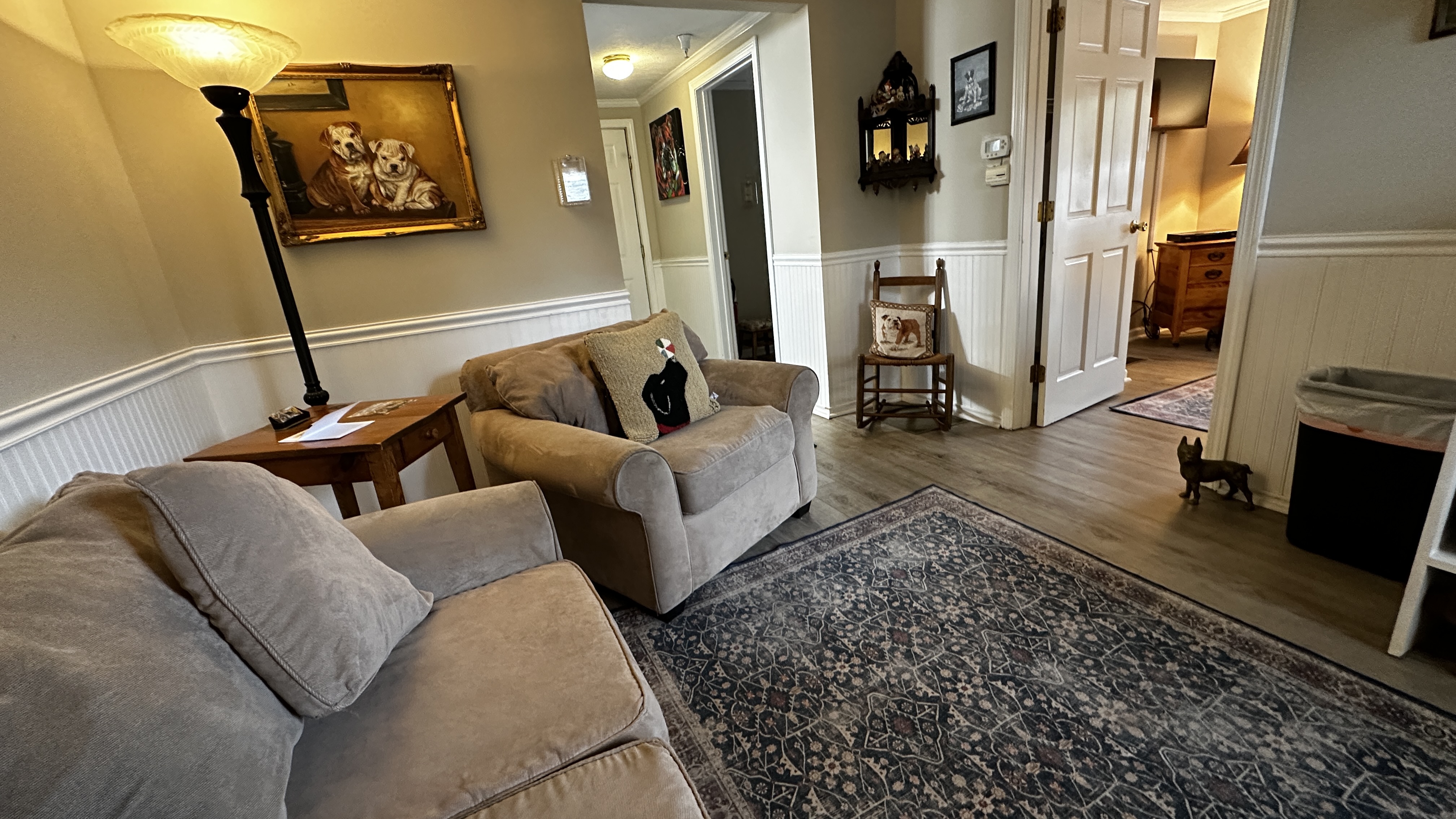 A living area featuring a light grey armchair with a black bear decorative pillow. The space includes a white storage unit equipped with a microwave, mini-fridge, and coffee maker, situated beneath a wall-mounted TV. In the background, doorways lead to other rooms and a hallway with a wooden rocking chair.