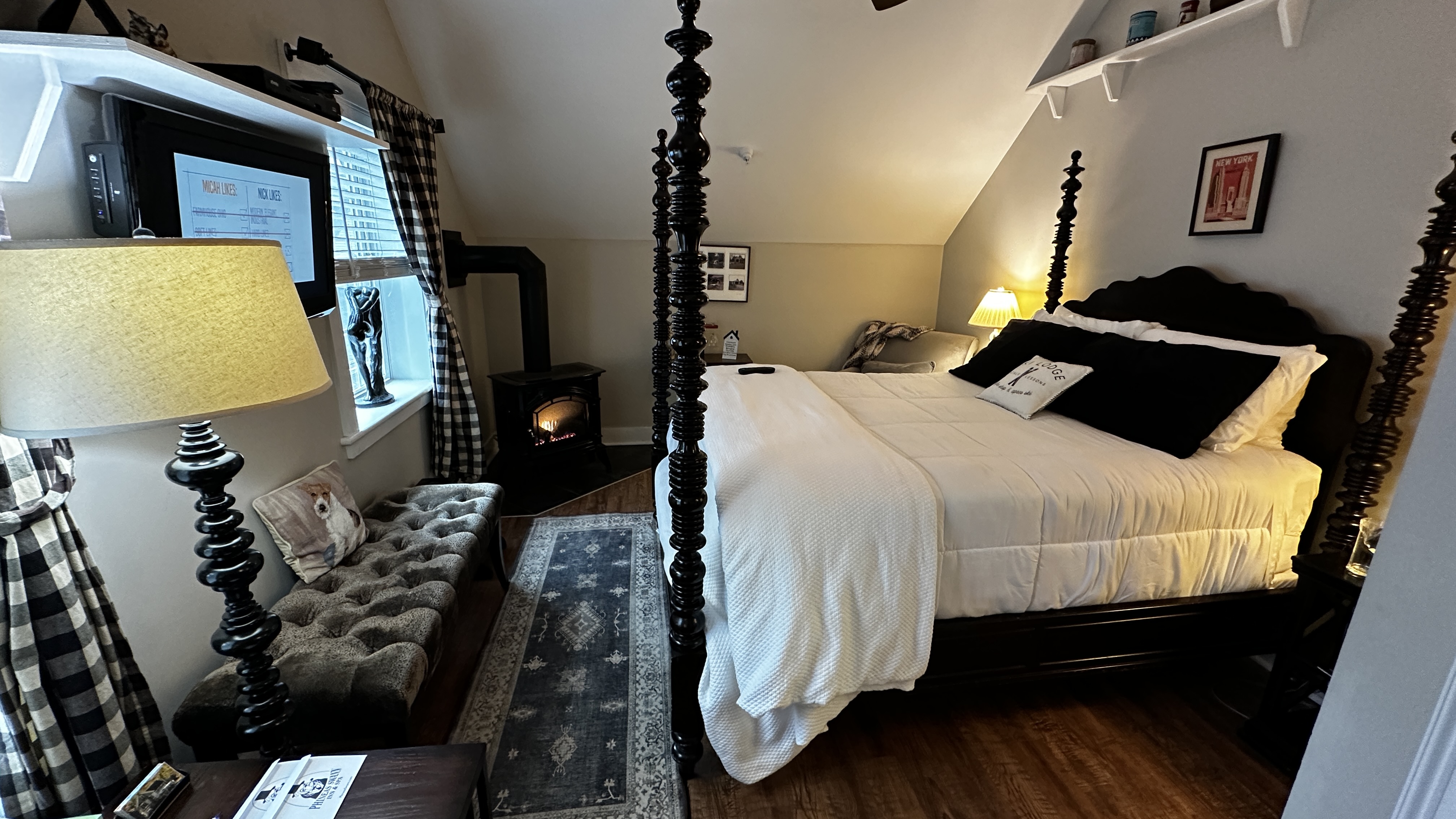 A cozy guest bedroom featuring a dark wood queen-size four-poster bed with crisp white linens and black accent pillows. The room includes a tufted gray bench, a gas-log fireplace, a wall-mounted TV, and sloped ceilings that create an intimate attic-suite atmosphere.