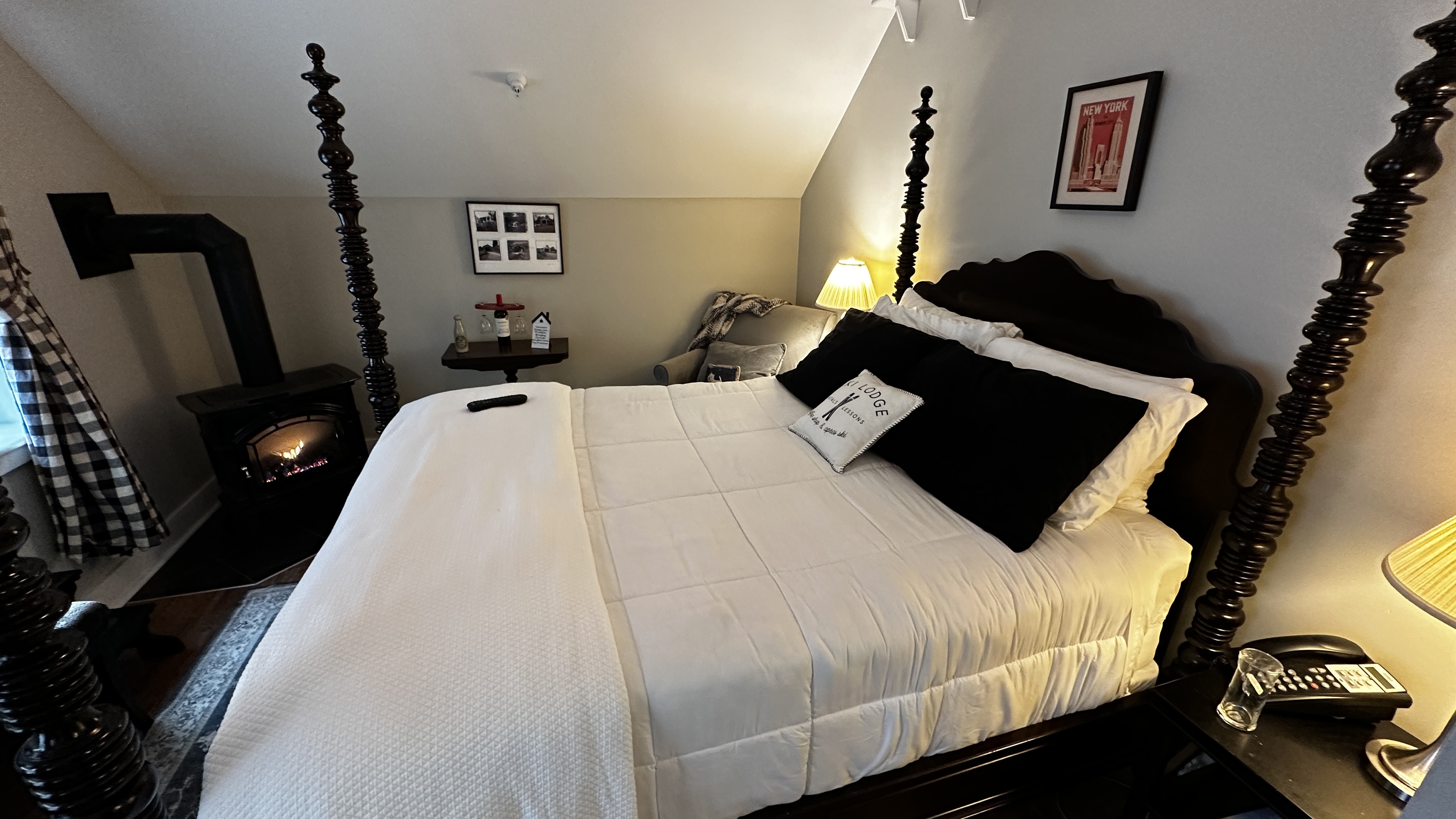 An attic-style guest room featuring a queen-size four-poster bed with crisp white linens and black accent pillows. The room includes a cozy gas-log fireplace, a tufted gray bench, and a wall-mounted TV, all set under a charming sloped ceiling.
