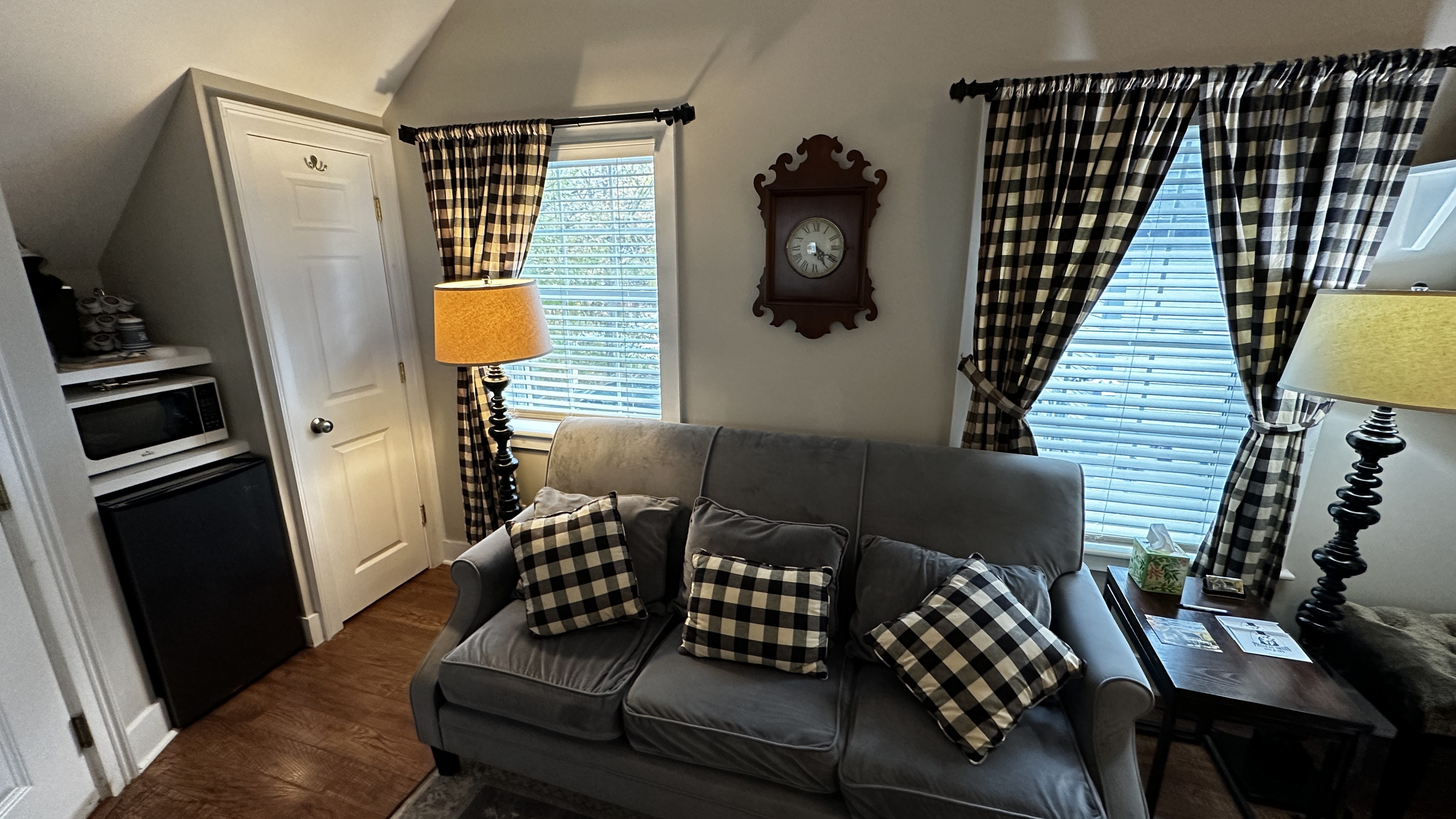 A cozy seating area in an attic-style suite, featuring a plush gray sofa with black and white checkered pillows. The space includes a small kitchenette nook with a microwave and mini-fridge, windows with checkered curtains, and a traditional wall clock.