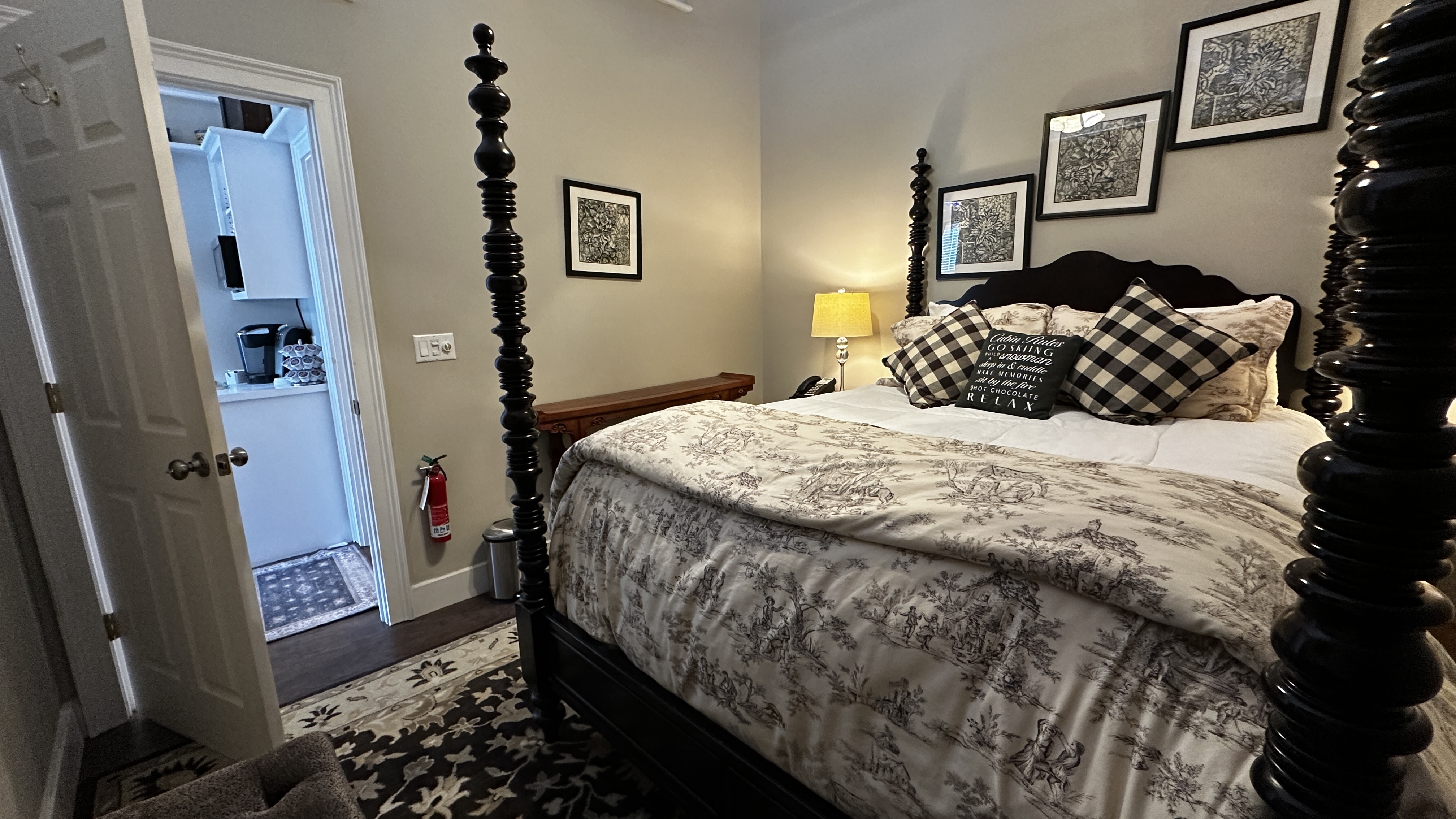 A cozy bedroom featuring a black four-poster bed with a patterned gray and white duvet and buffalo plaid accent pillows. The room includes several framed black-and-white photos on the wall, a bedside lamp casting a warm glow, and an open doorway leading to a brightly lit kitchenette area.