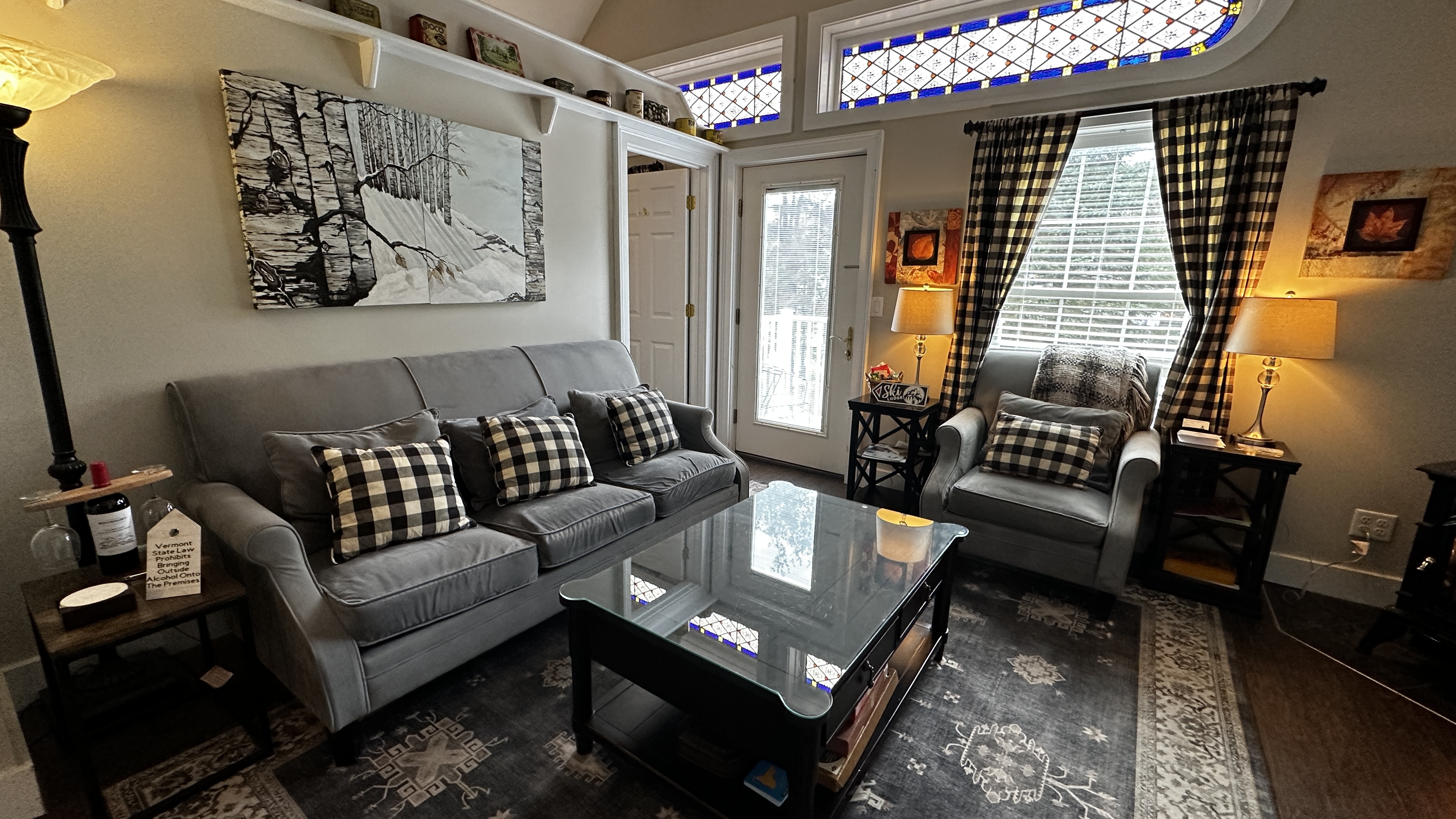 A farmhouse-style living room featuring a long gray sofa with buffalo plaid pillows and a glass-top coffee table on a large patterned area rug. The room is decorated with a large winter-themed wall art piece, warm lamps, and a unique stained-glass transom window above a white door leading to an outdoor space.