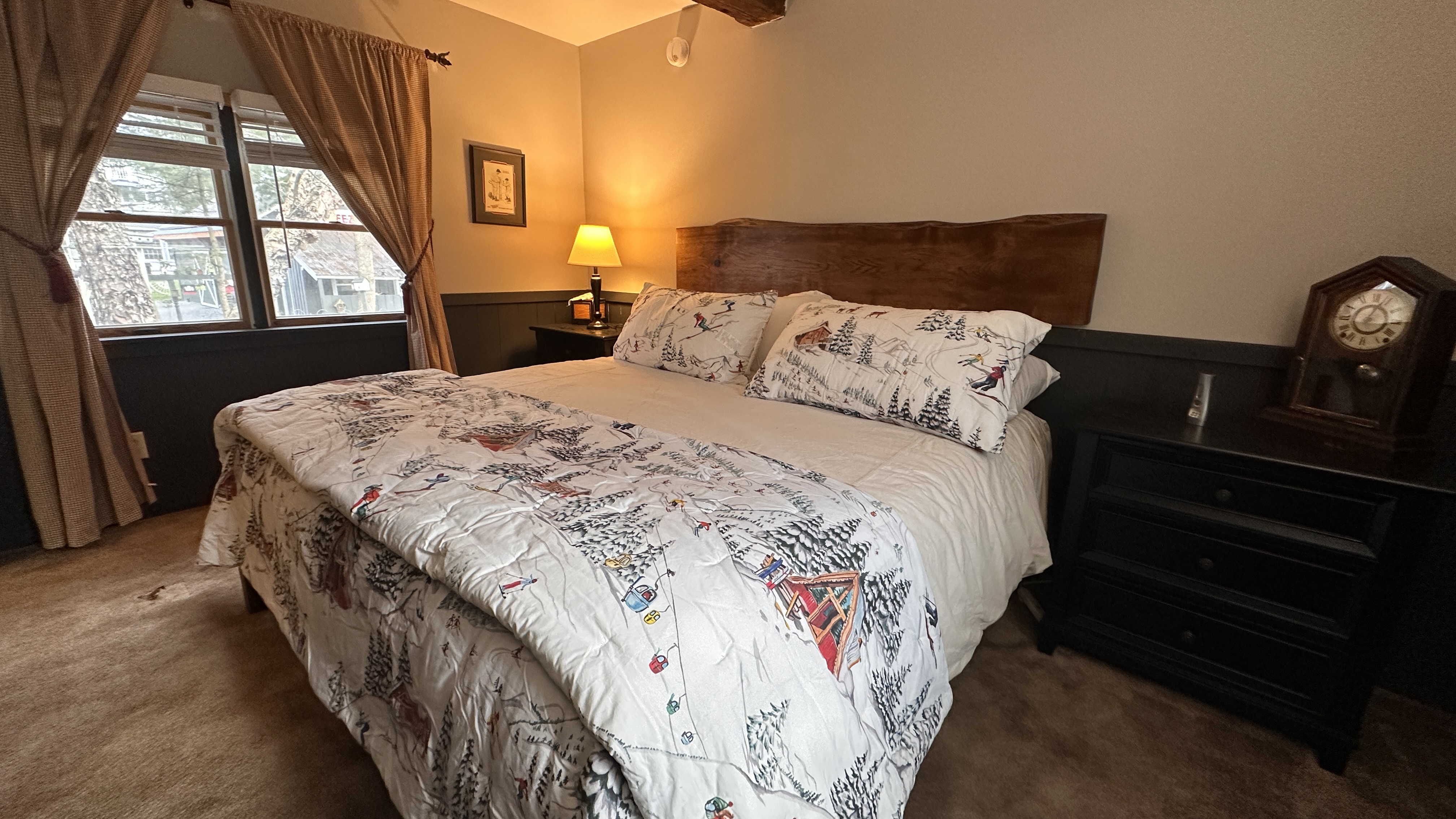 A cozy bedroom featuring a wooden king-sized bed with a live-edge headboard and white bedding with a winter-themed pattern. The room includes a black bedside table with a vintage-style clock, a warm lamp, and a large window with tan curtains overlooking a wooded area.