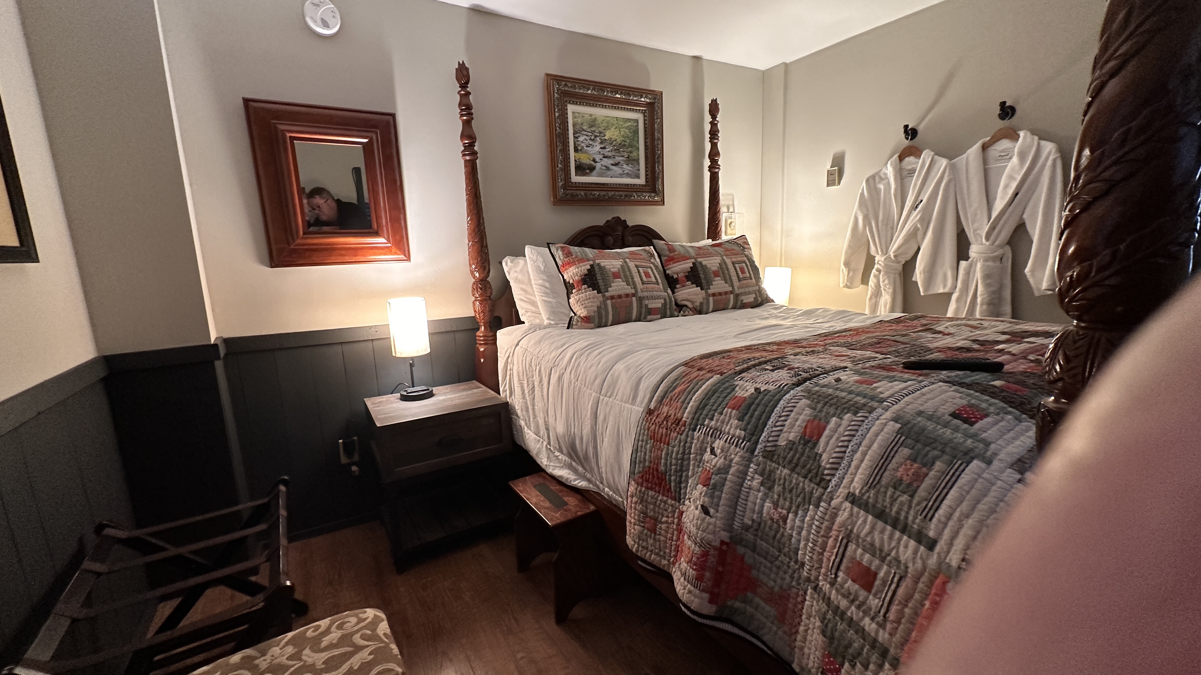 A cozy bedroom featuring a dark wood four-poster bed dressed in white linens and a red and gray patterned quilt. The room includes dark-paneled wainscoting, a bedside nightstand with a warm glowing lamp, and white plush robes hanging on the wall.