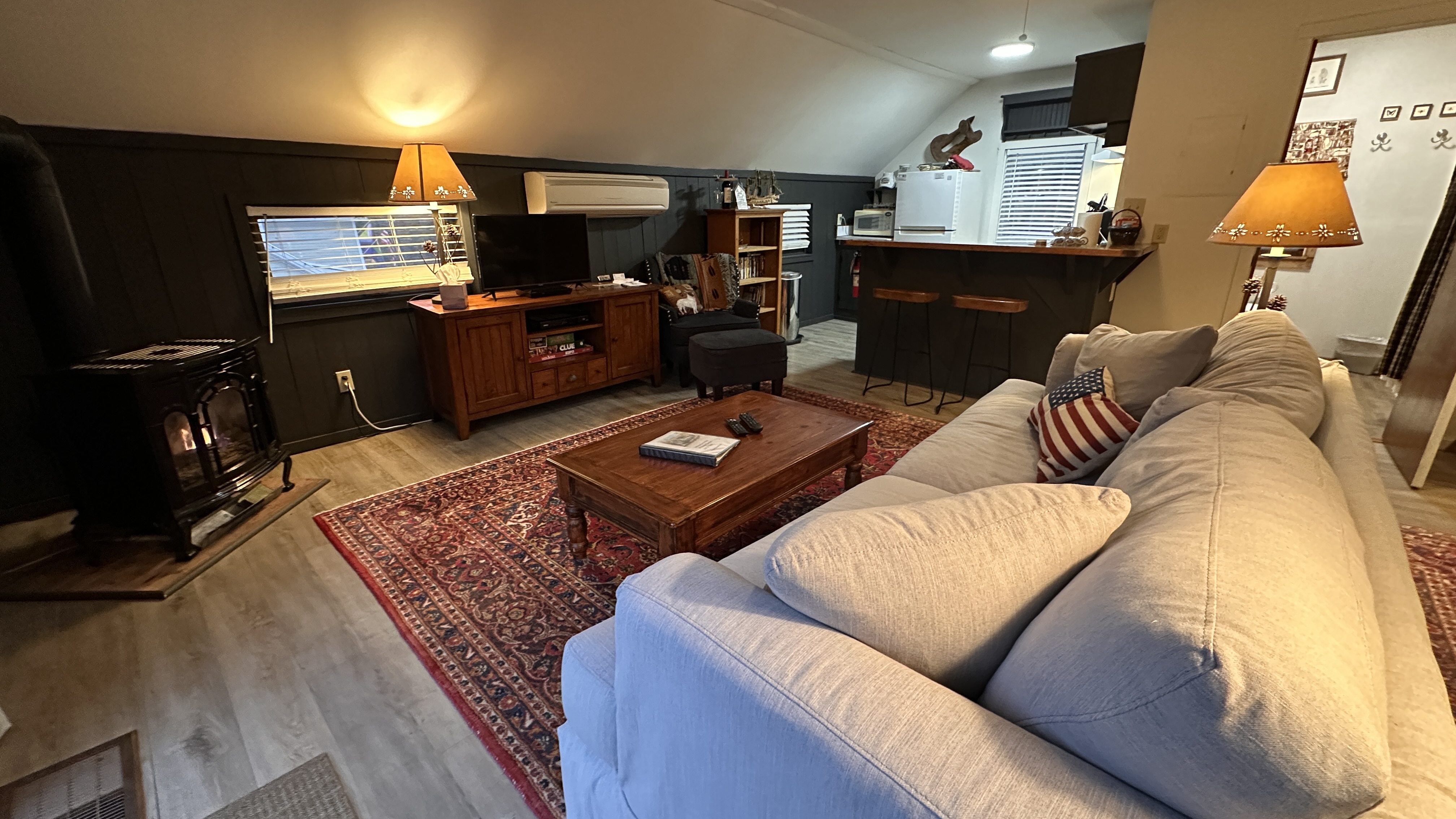 A cozy attic-style living area featuring a light-colored sofa facing a wooden coffee table on a red patterned rug, with a fireplace in the corner and a breakfast bar leading into a small kitchen in the background.