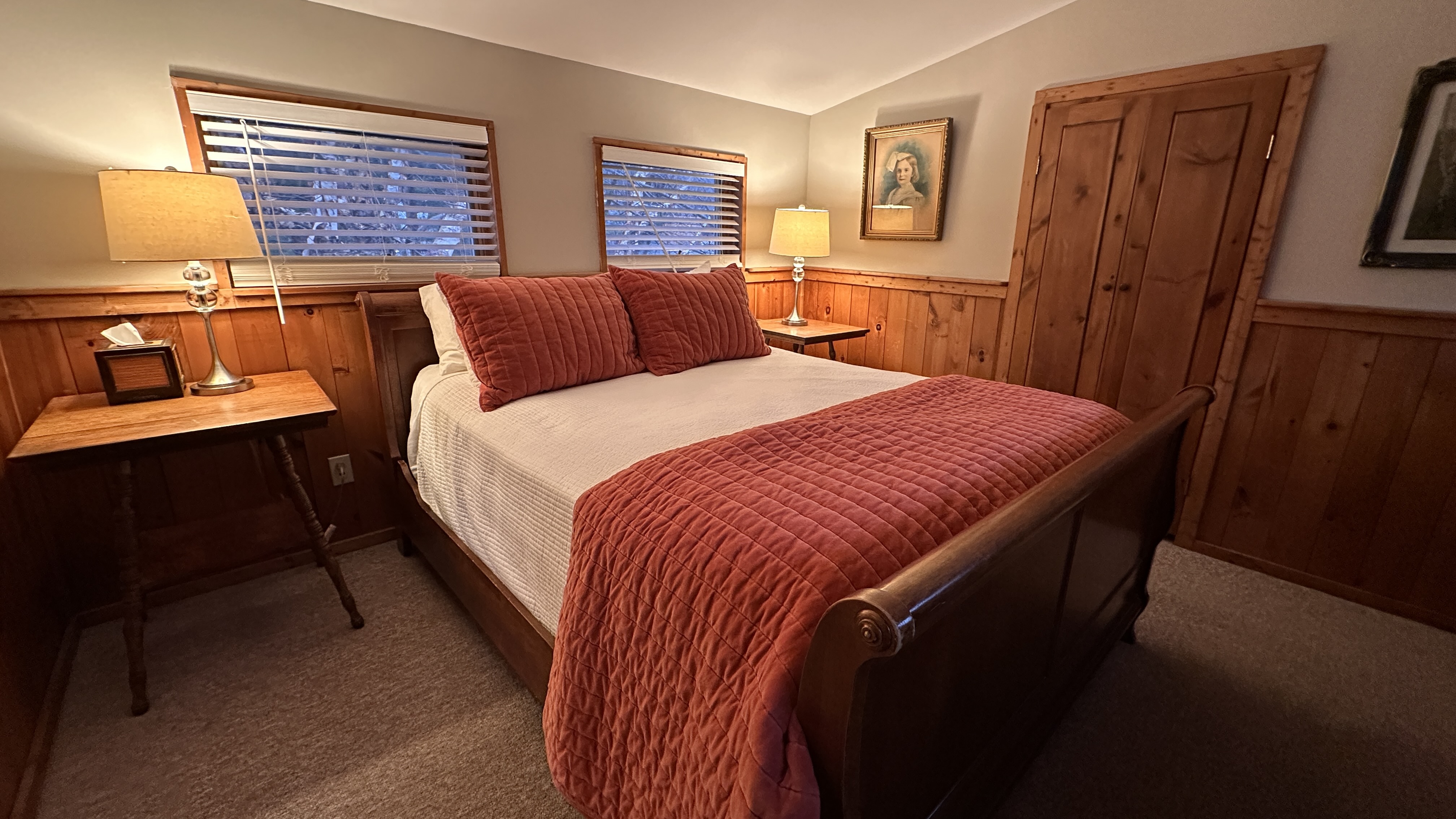 A partially wood-paneled bedroom with a sleigh-style bed featuring a white comforter and reddish-orange textured pillows and throw, flanked by small wooden nightstands with lamps.