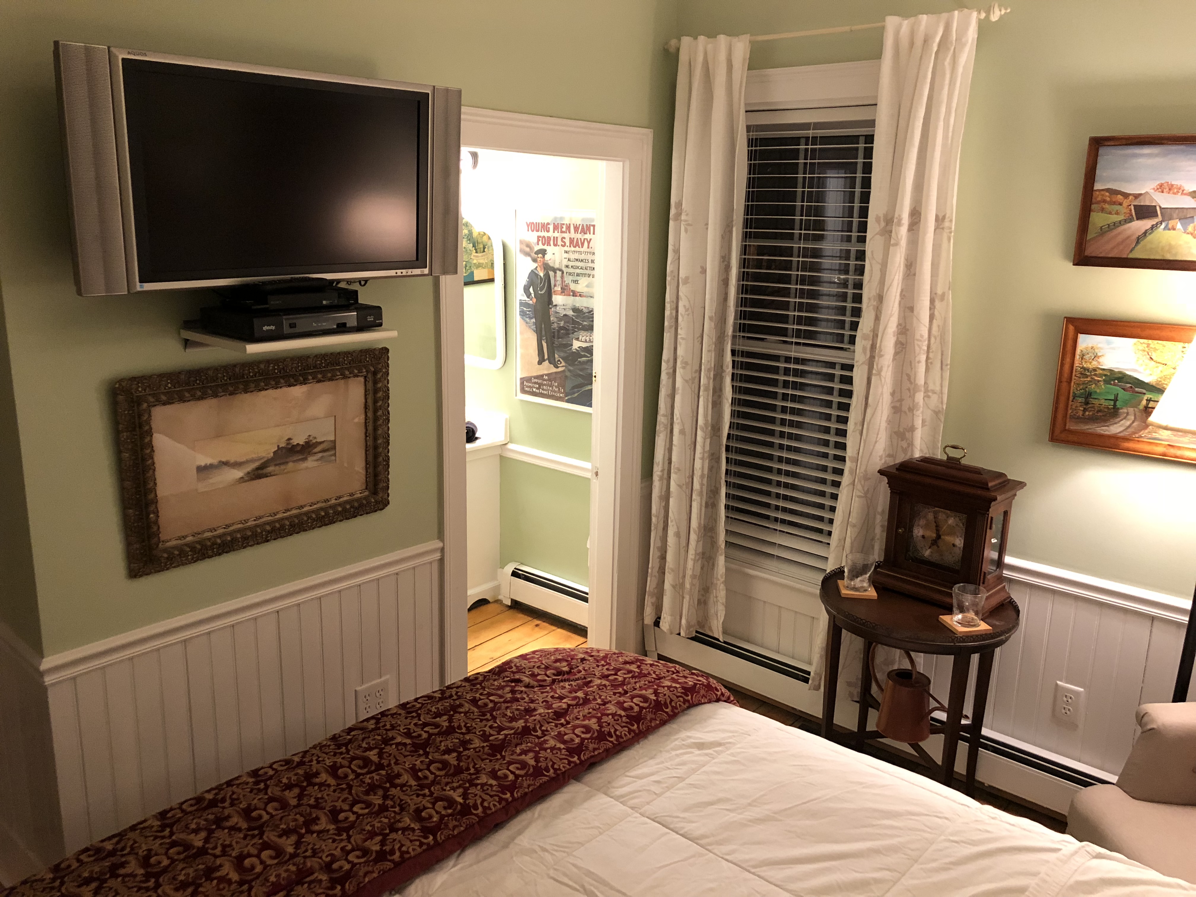 A bedroom with light green walls and white wainscoting. A television is mounted on the wall above a framed picture, and a doorway leads into an adjacent room. The space also features a window with white curtains, a small wooden table holding a vintage-style clock, and the foot of a bed with a dark patterned throw.