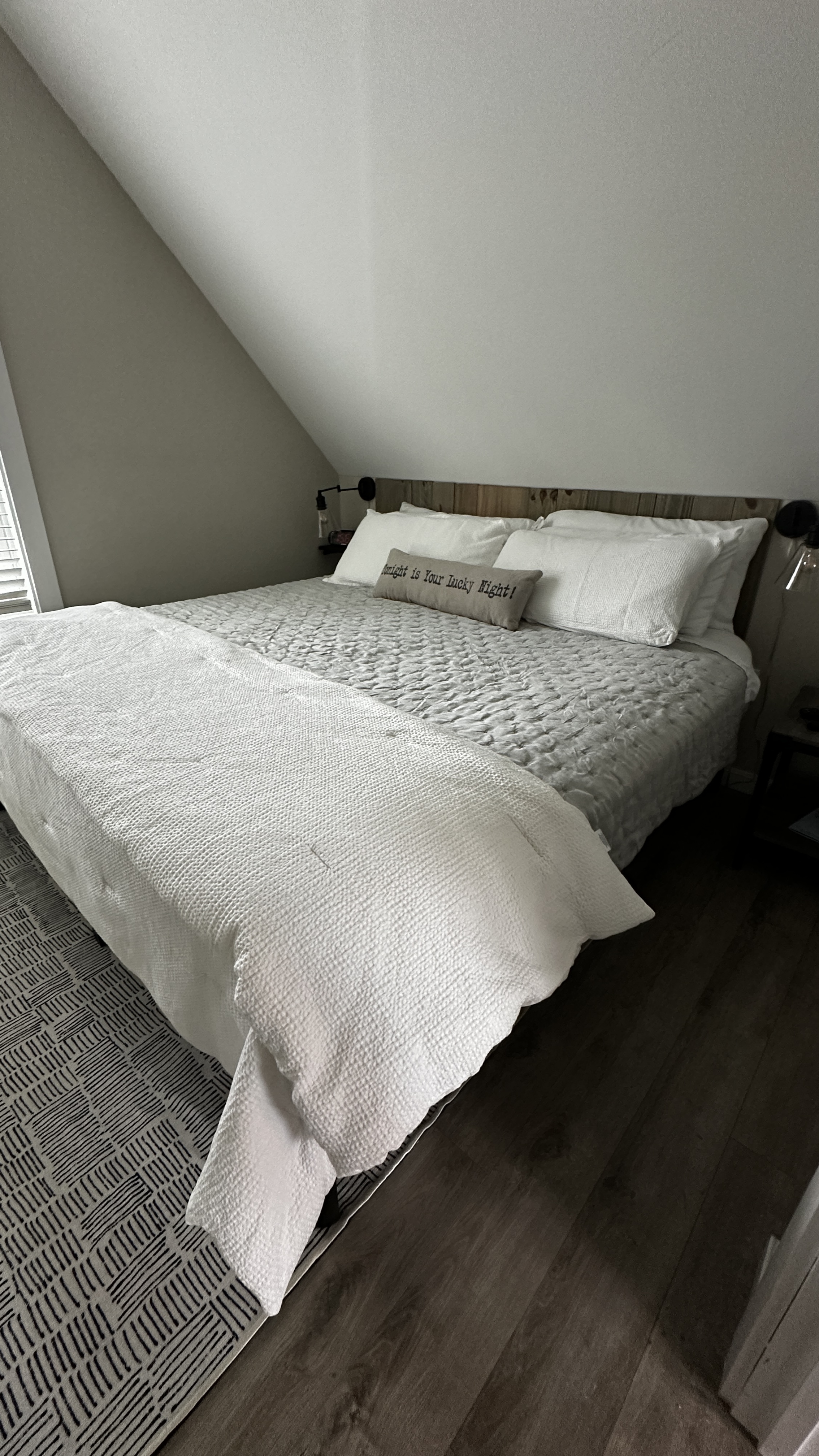 A close-up of a bed with a grey textured comforter and a white folded blanket at the foot. The bed features white pillows, a long grey accent pillow, and a rustic wooden headboard, set against a white slanted wall in an attic-style room.