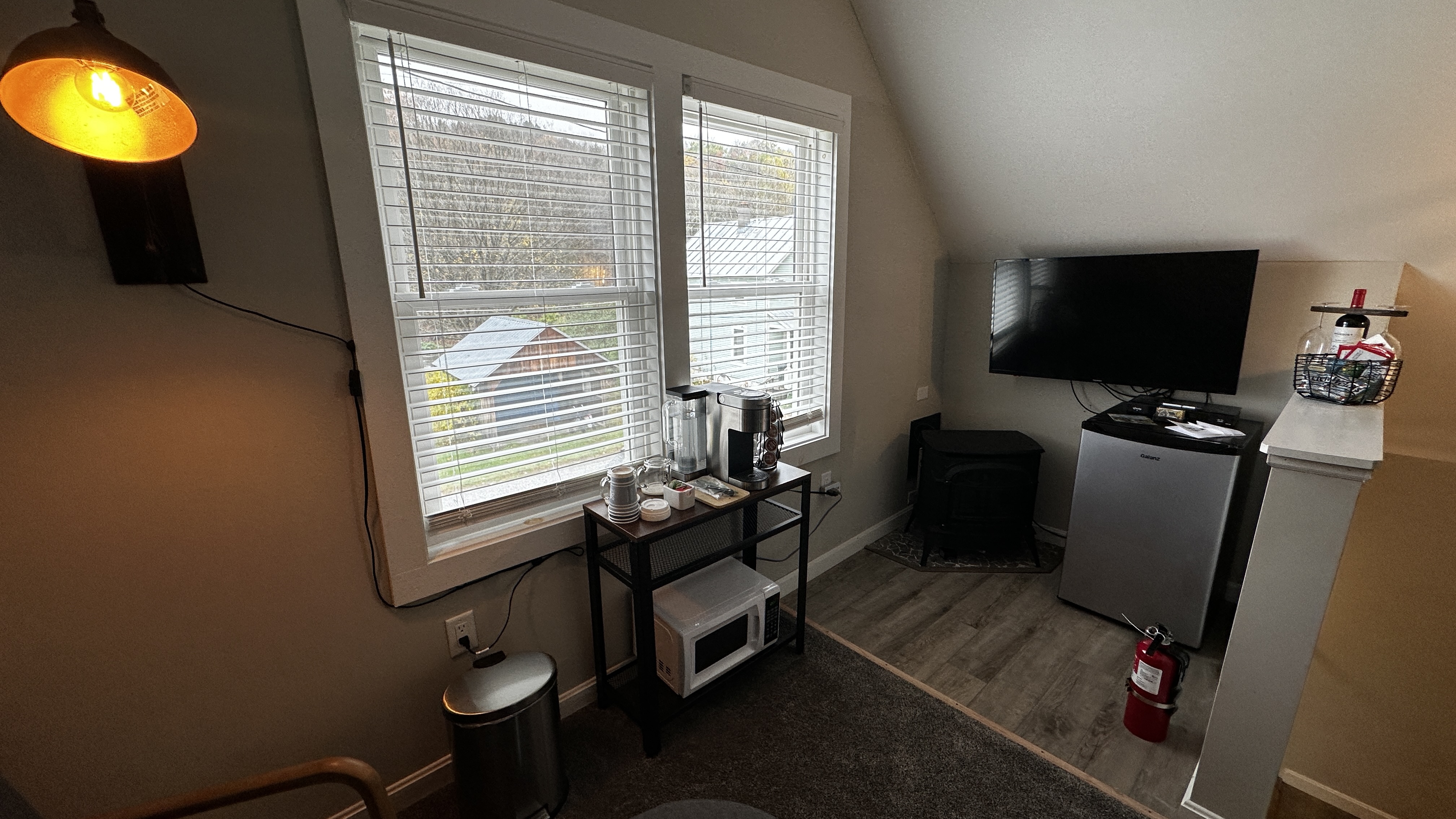 A compact kitchenette nook in an attic-style room with a slanted ceiling. The space features two windows with blinds, a small black table holding a coffee maker and supplies, a white microwave on the lower shelf, and a mini-fridge tucked into a corner under a wall-mounted television.