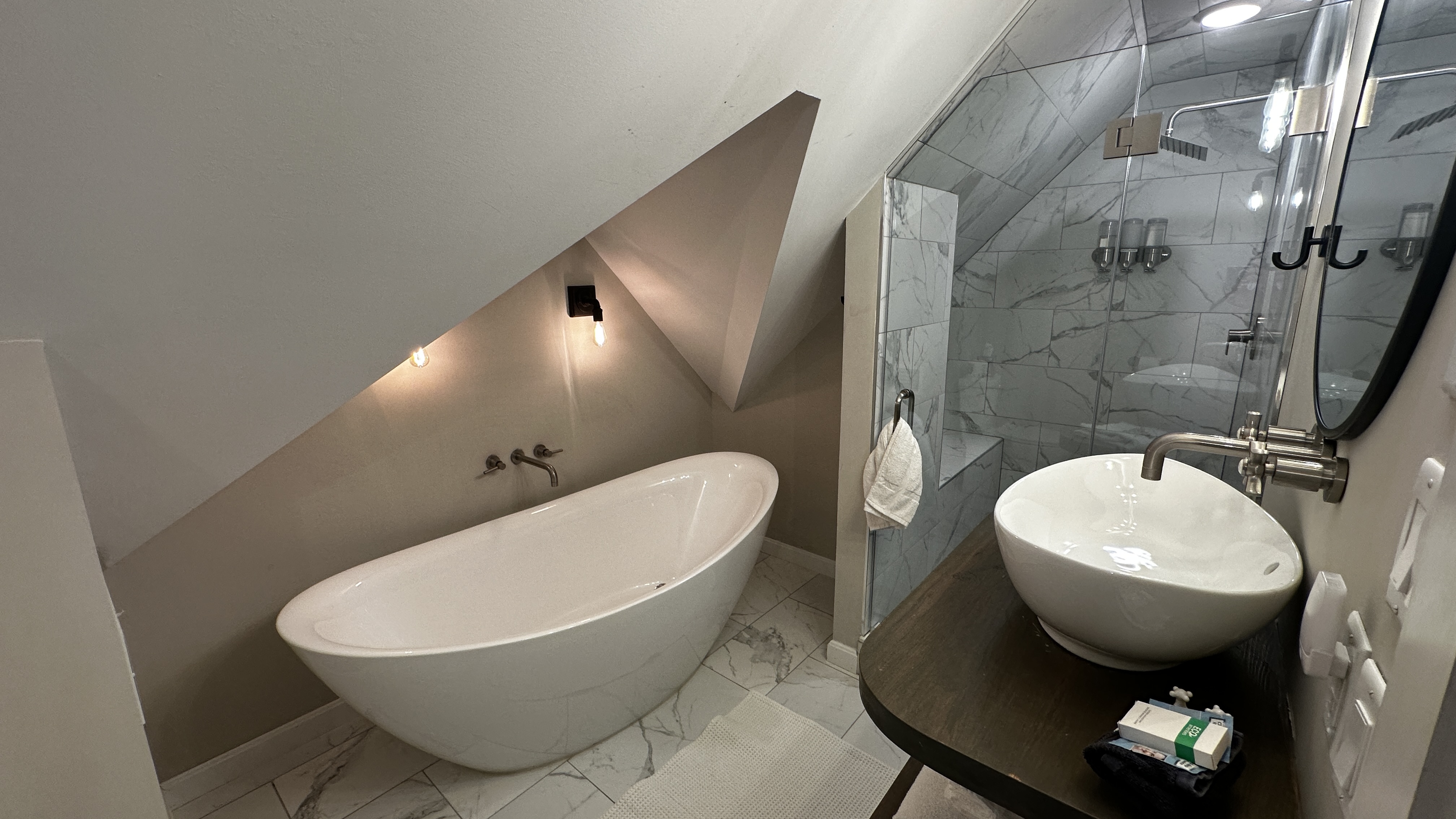 A luxurious, modern bathroom tucked into an attic space with a sharply slanted white ceiling. The room features a large, oval-shaped freestanding soaking tub and a sleek vessel sink on a dark wood vanity. To the right, a glass-enclosed walk-in shower with marble-patterned tile is visible.