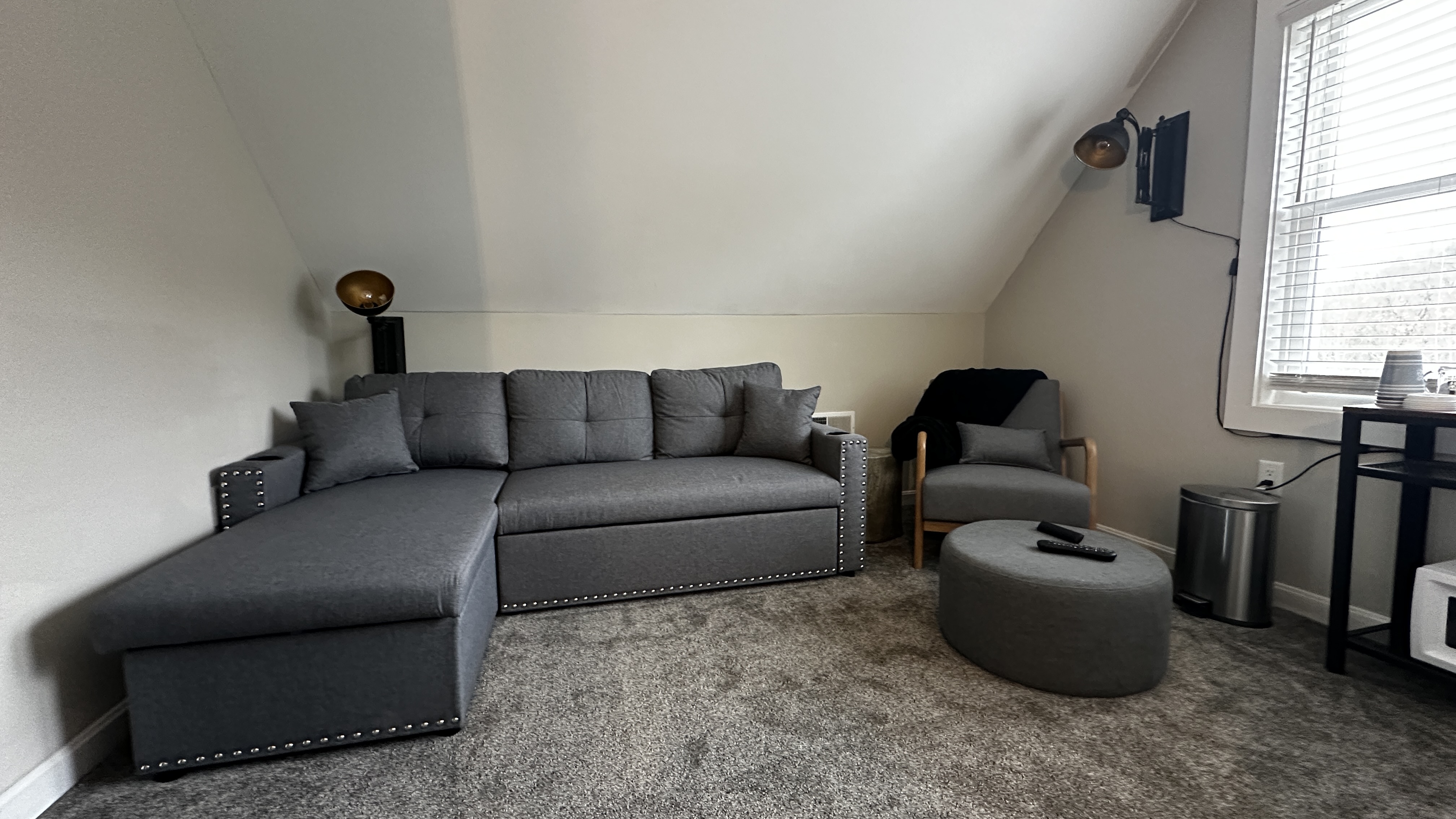 A spacious living area in an attic suite featuring a dark grey sectional sofa and a matching armchair on a plush grey carpet. To the right, a small black utility table sits under two bright windows, and the room is illuminated by warm, modern wall lamps against a slanted white ceiling.