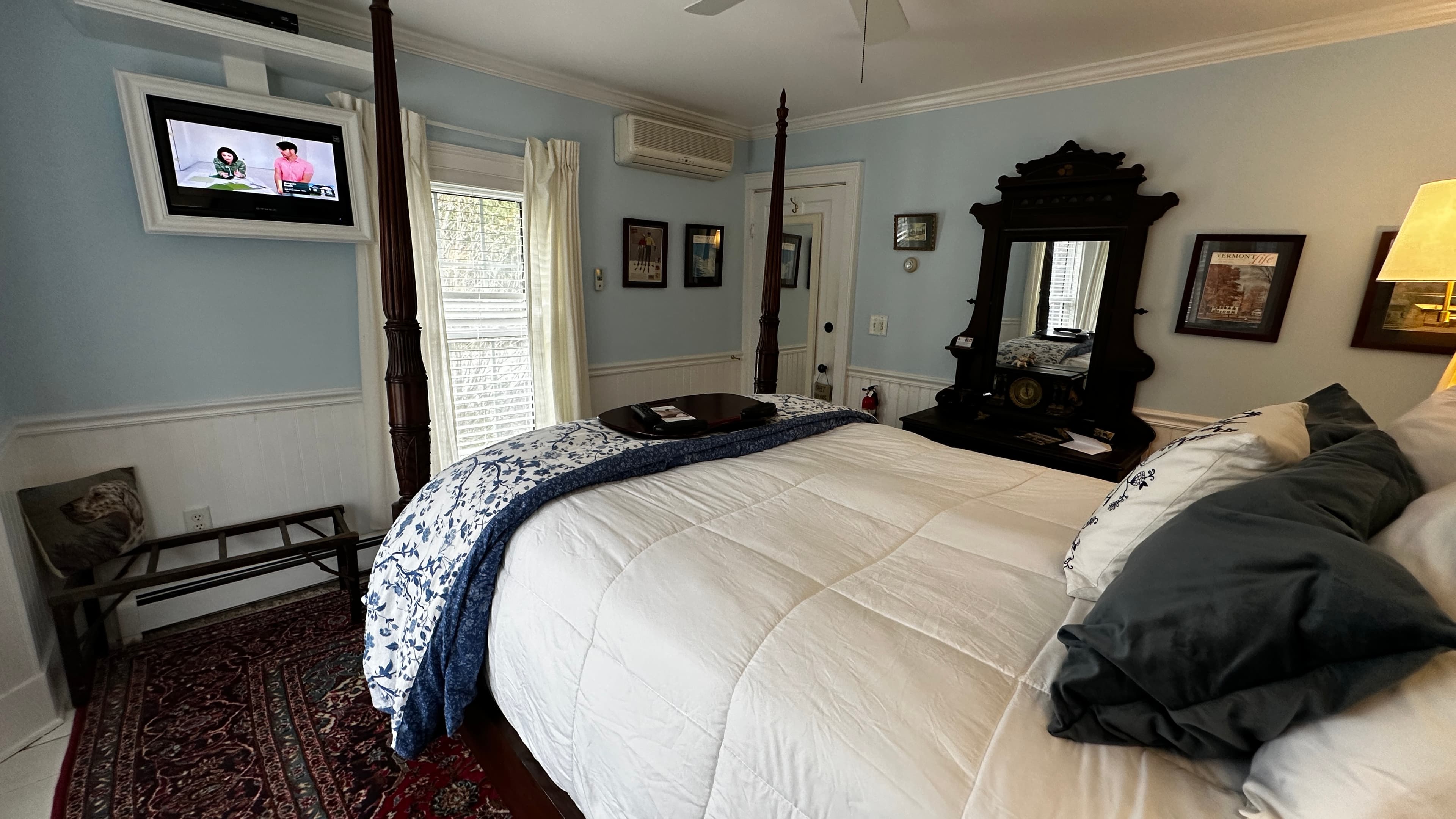 A dark wood four-poster bed with white and blue patterned bedding, an ornate antique dresser with a mirror, a wall-mounted TV, and light blue walls with white wainscoting.