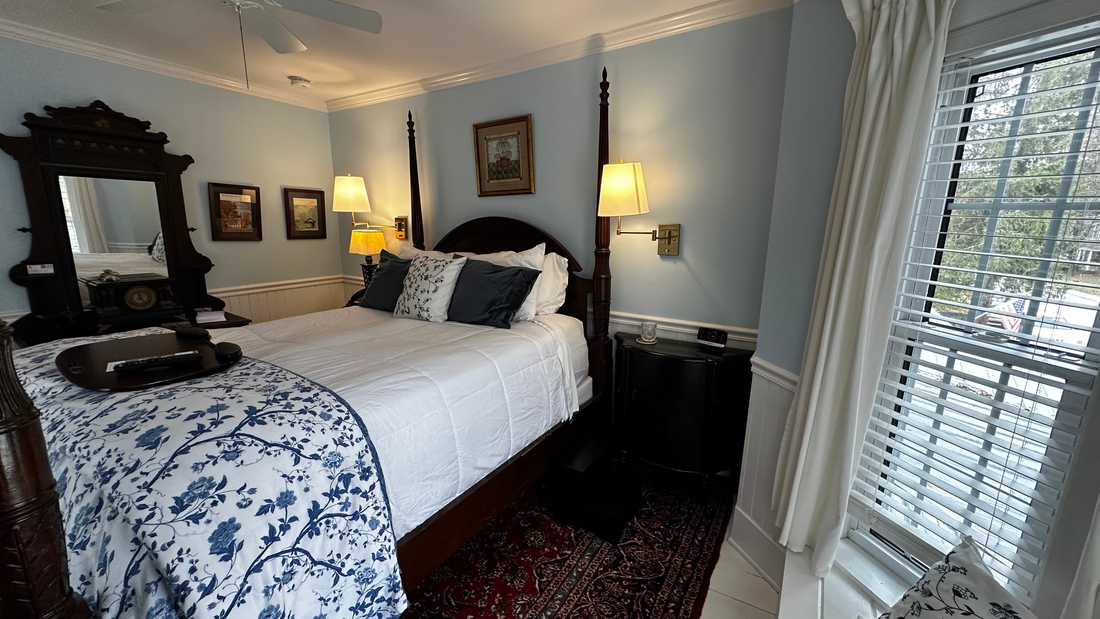 An antique dark wood dresser with a large, ornate mirror stands against light blue walls in the Blue Room. The shot also features a corner of the four-poster bed and a traditional red patterned rug.