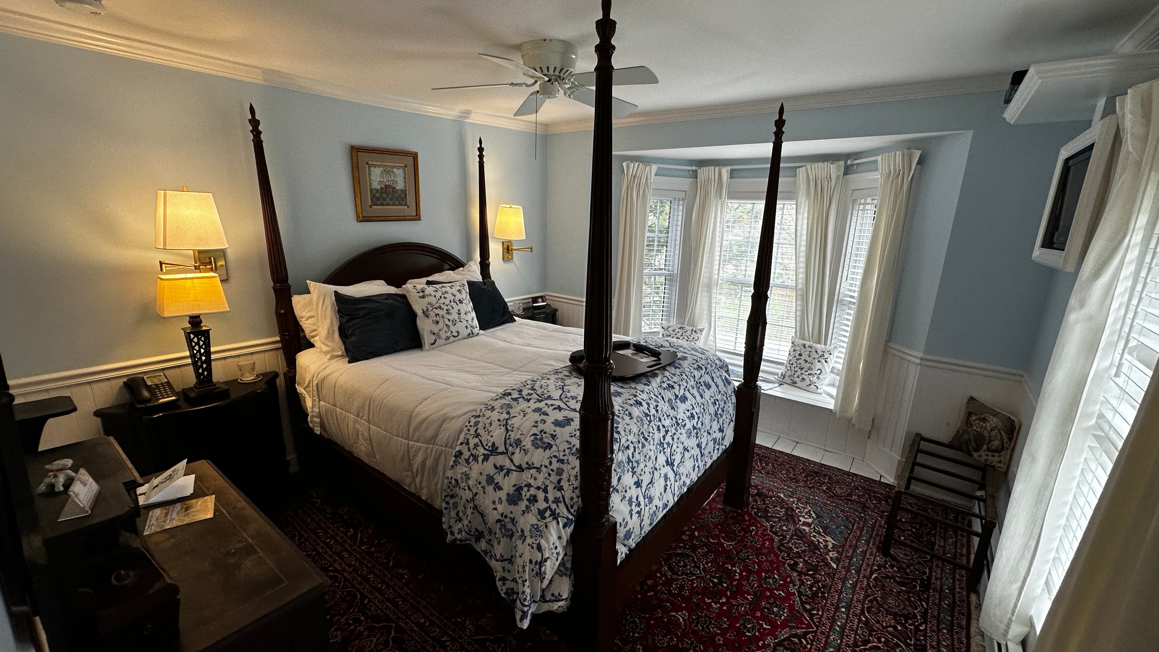 A traditional guest room with light blue walls, featuring a dark wood four-poster bed with blue and white floral bedding, a large bay window with a window seat, and a red patterned rug.