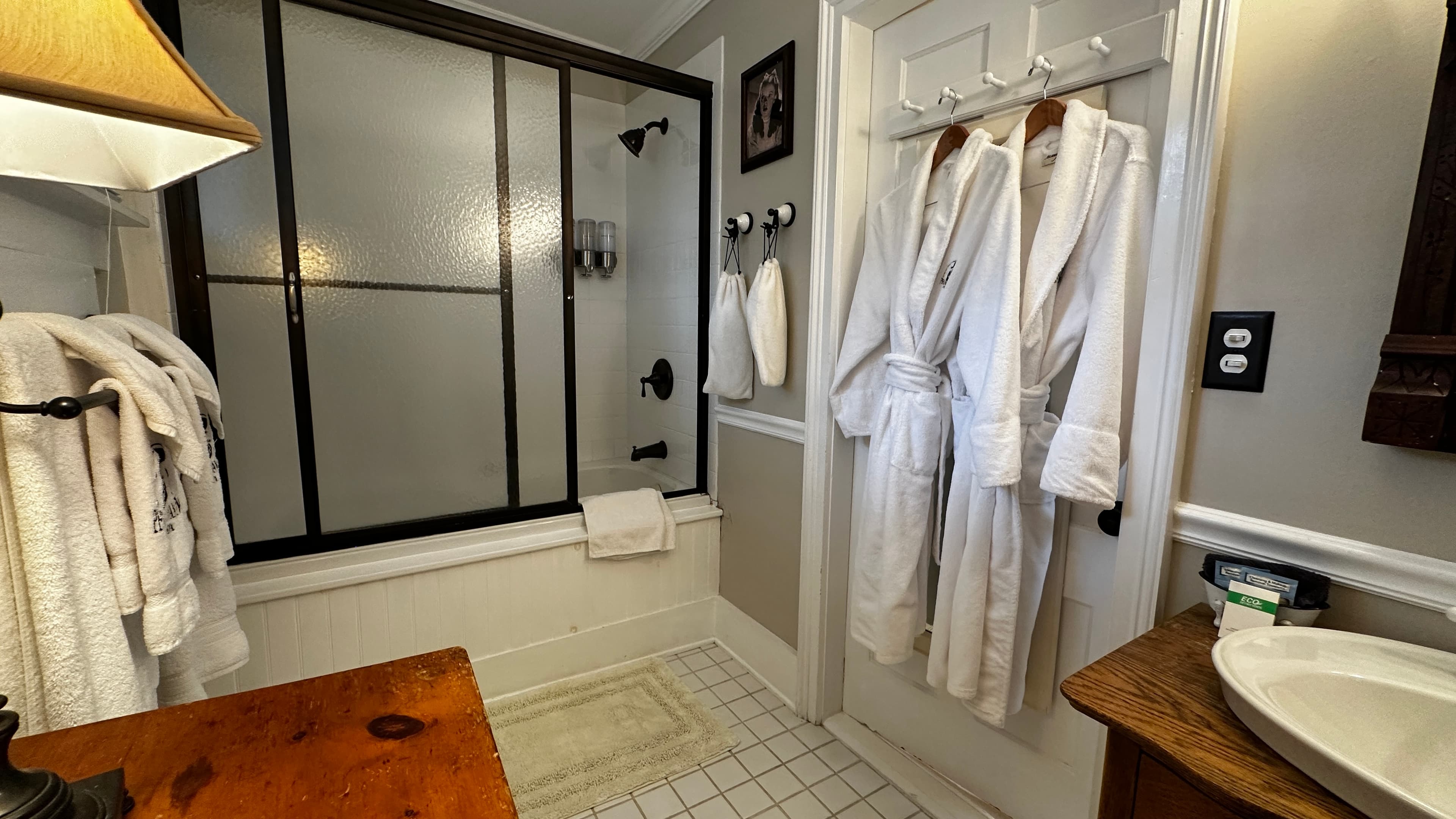 A modern bathroom with a white vessel sink on a wooden vanity, two white robes hanging on a white door, and a bathtub with a black-framed sliding glass door.