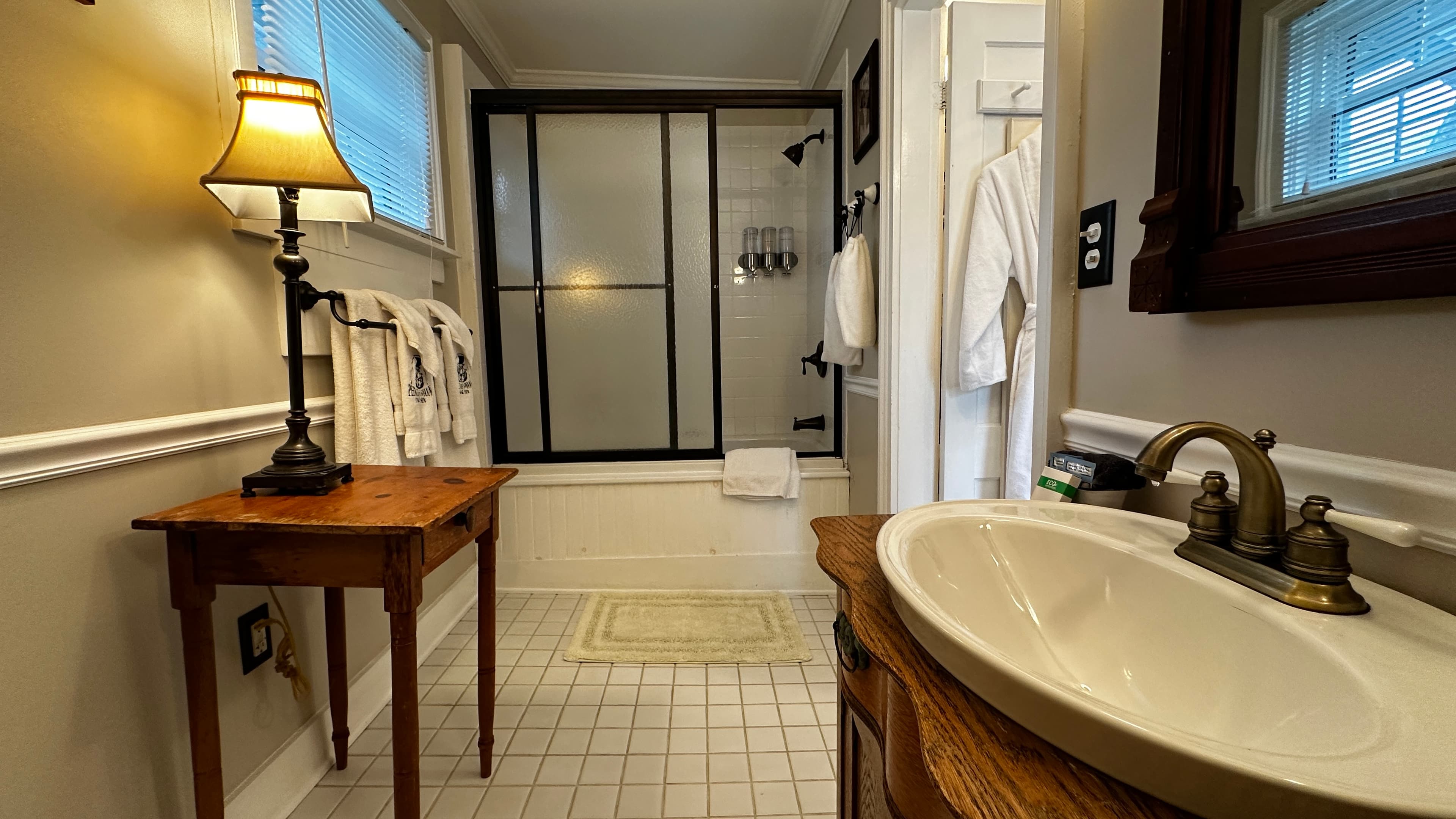 A classic bathroom featuring a white vessel sink on a rich wood vanity, an antique side table with a lamp, and a bathtub enclosed by black-framed frosted glass doors.