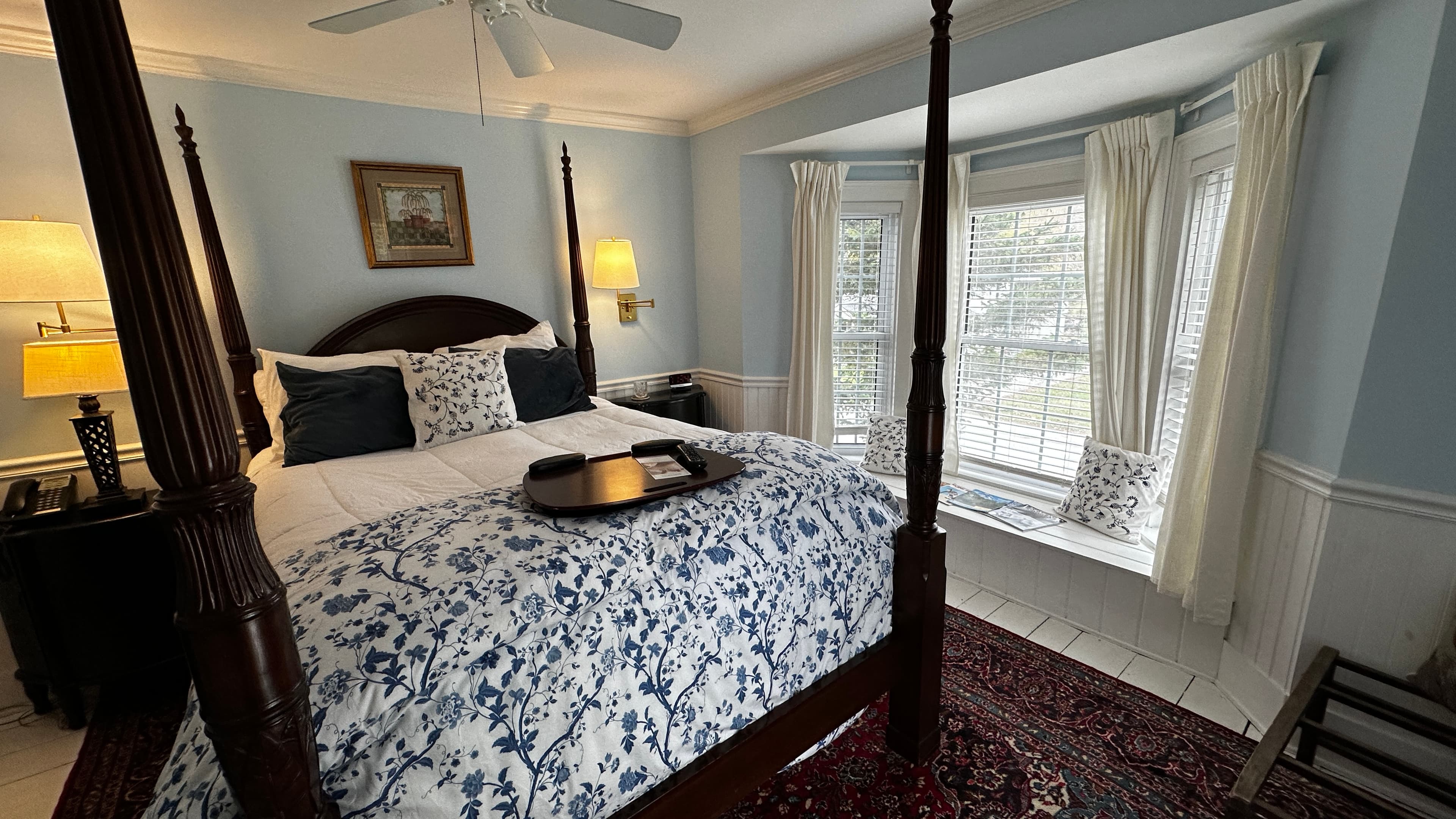 A bright guest room featuring light blue walls and a large bay window with a built-in window seat. A dark wood four-poster bed is dressed in blue and white floral bedding, set atop a decorative patterned rug.