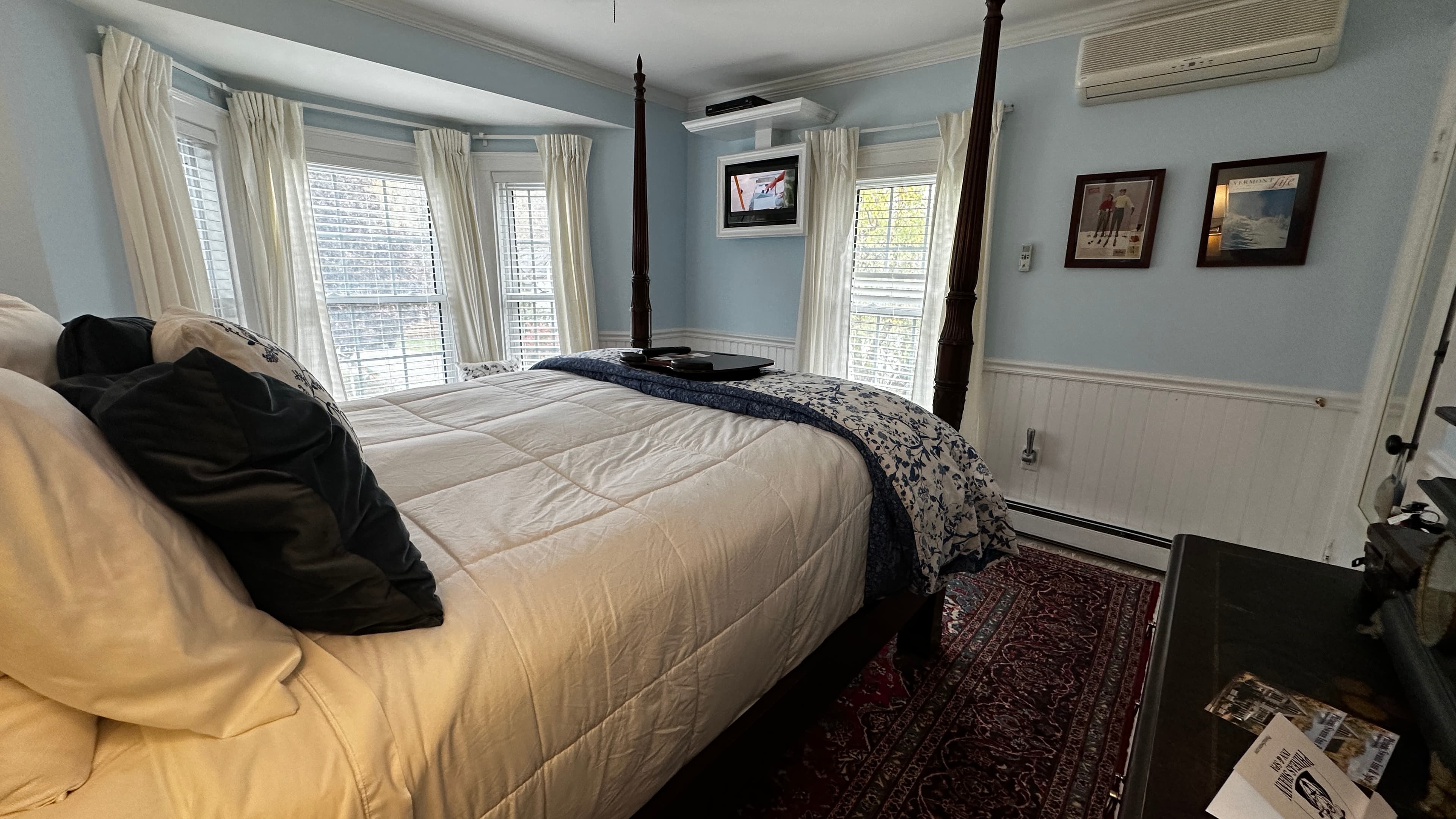 A dark wood four-poster queen bed with white and blue floral linens, set against light blue walls and a large bay window with a built-in window seat.