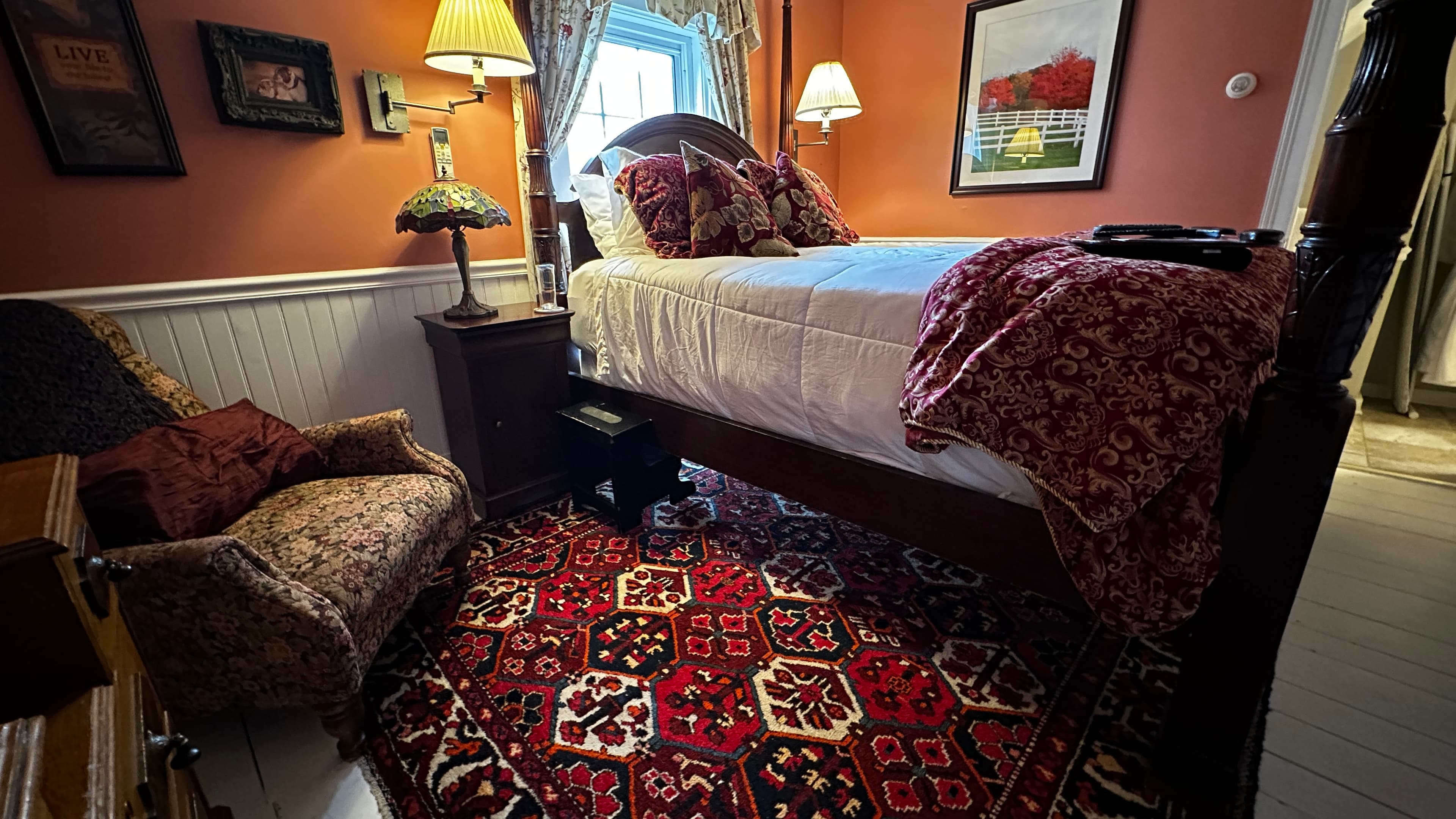 An elegant guest room featuring burnt sienna red walls, white wainscoting, and a dark wood four-poster queen bed. A patterned armchair sits beside the bed on a traditional red area rug, creating a warm and classic atmosphere.