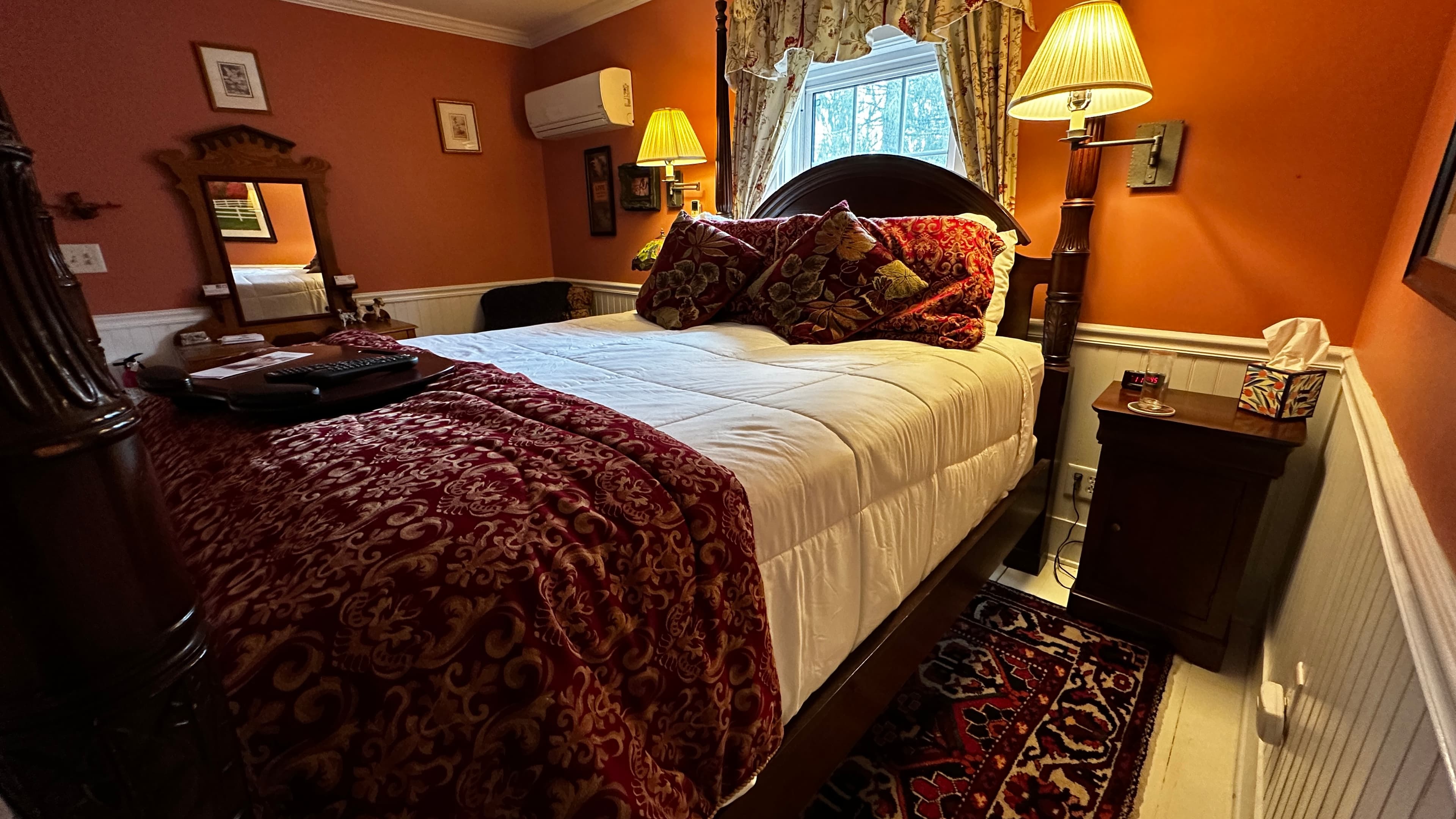 A cozy guest room with burnt sienna red walls and white wainscoting, featuring a dark wood queen-size four-poster bed with white linens and a red patterned throw. An antique dresser with a mirror and a bedside lamp add to the warm, traditional aesthetic.
