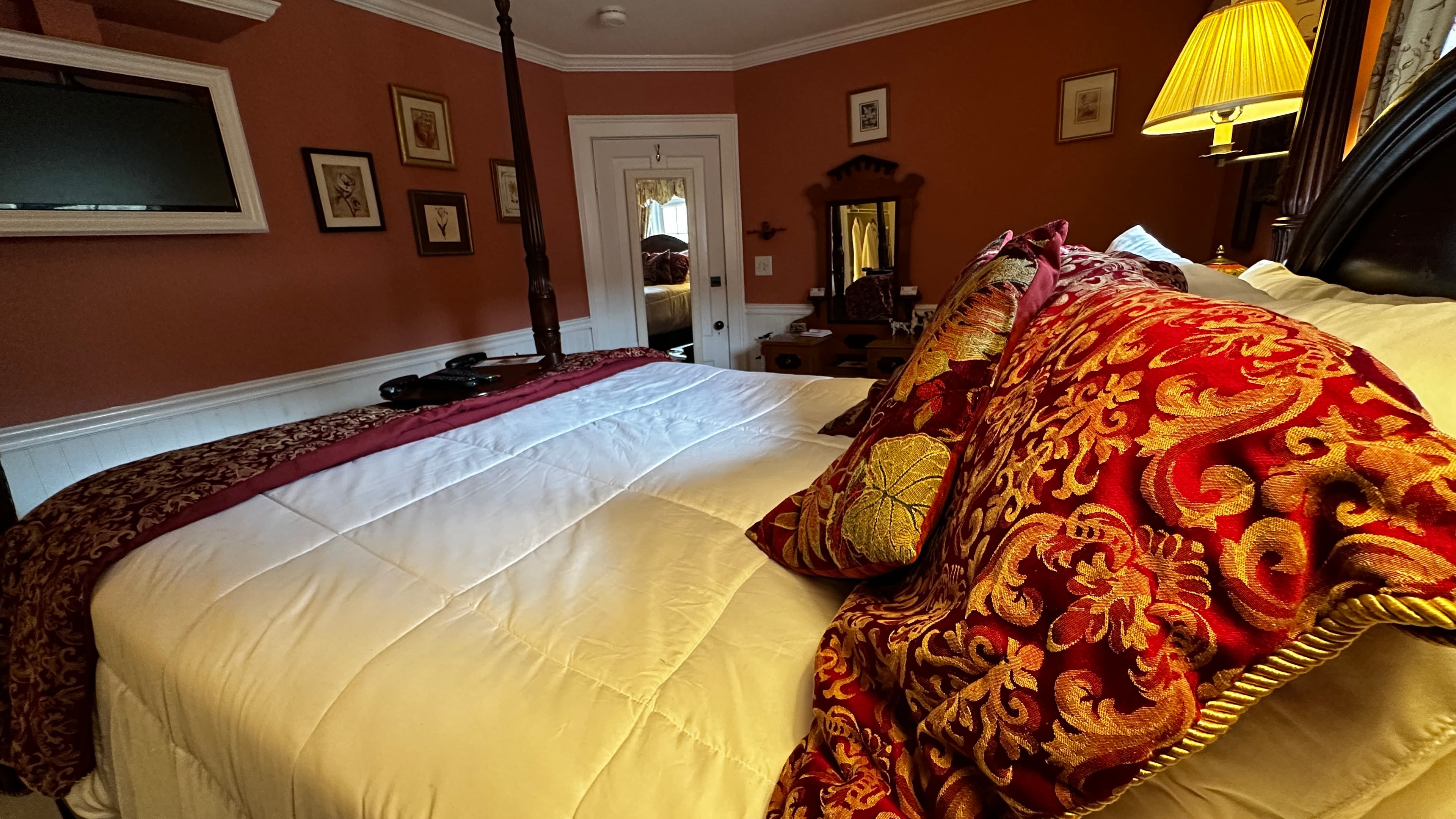 A guestroom with a dark wood four-poster bed with crisp white linens and a decorative red patterned throw. In the background, an antique wood dresser with an ornate mirror sits against burnt sienna walls with white wainscoting.