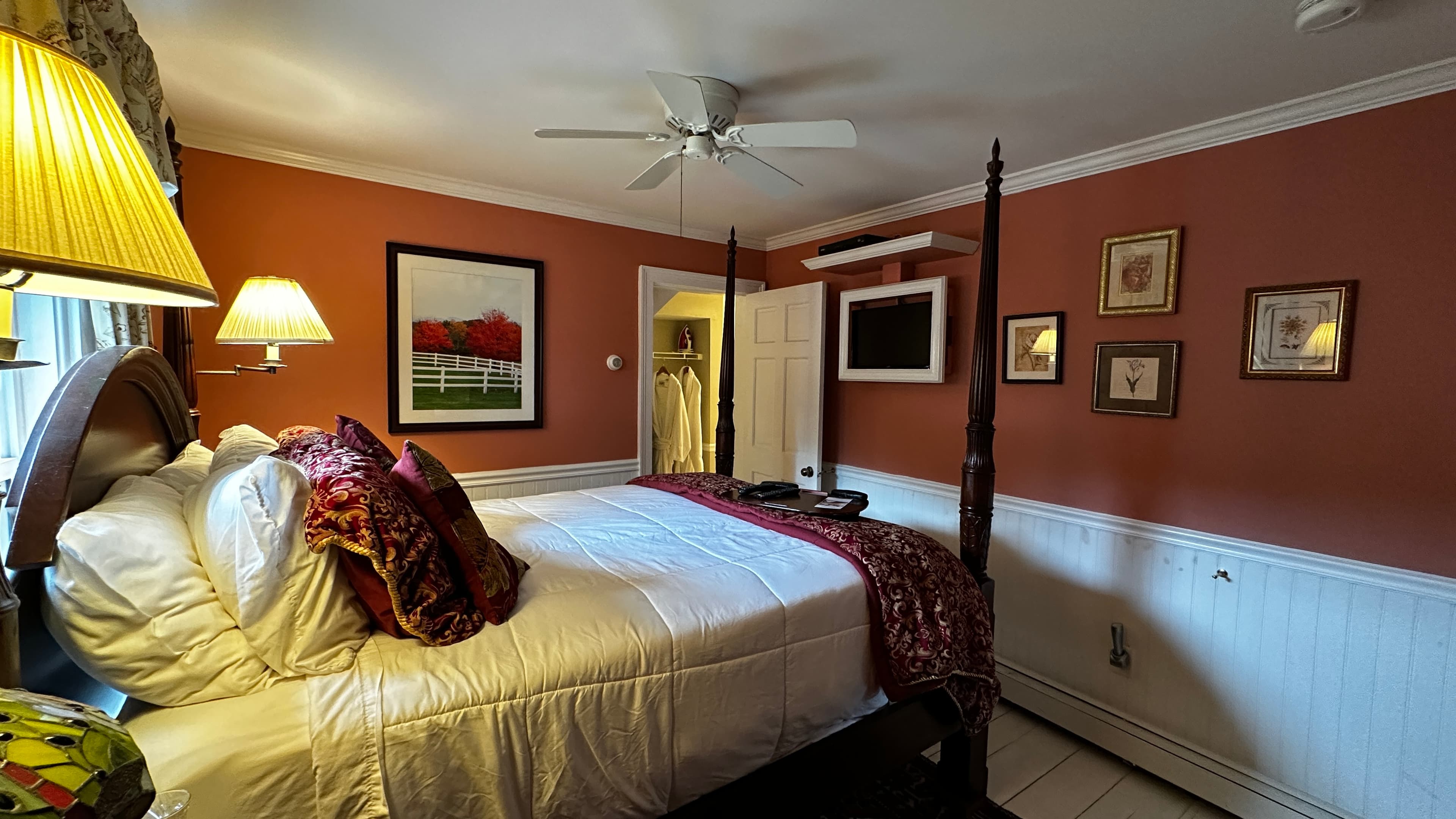 Red Room guest room with a dark wood queen-size four-poster bed with crisp white linens and a decorative red patterned throw. The room features burnt sienna walls above white beaded wainscoting, several framed pictures, and a wall-mounted flat-screen TV.