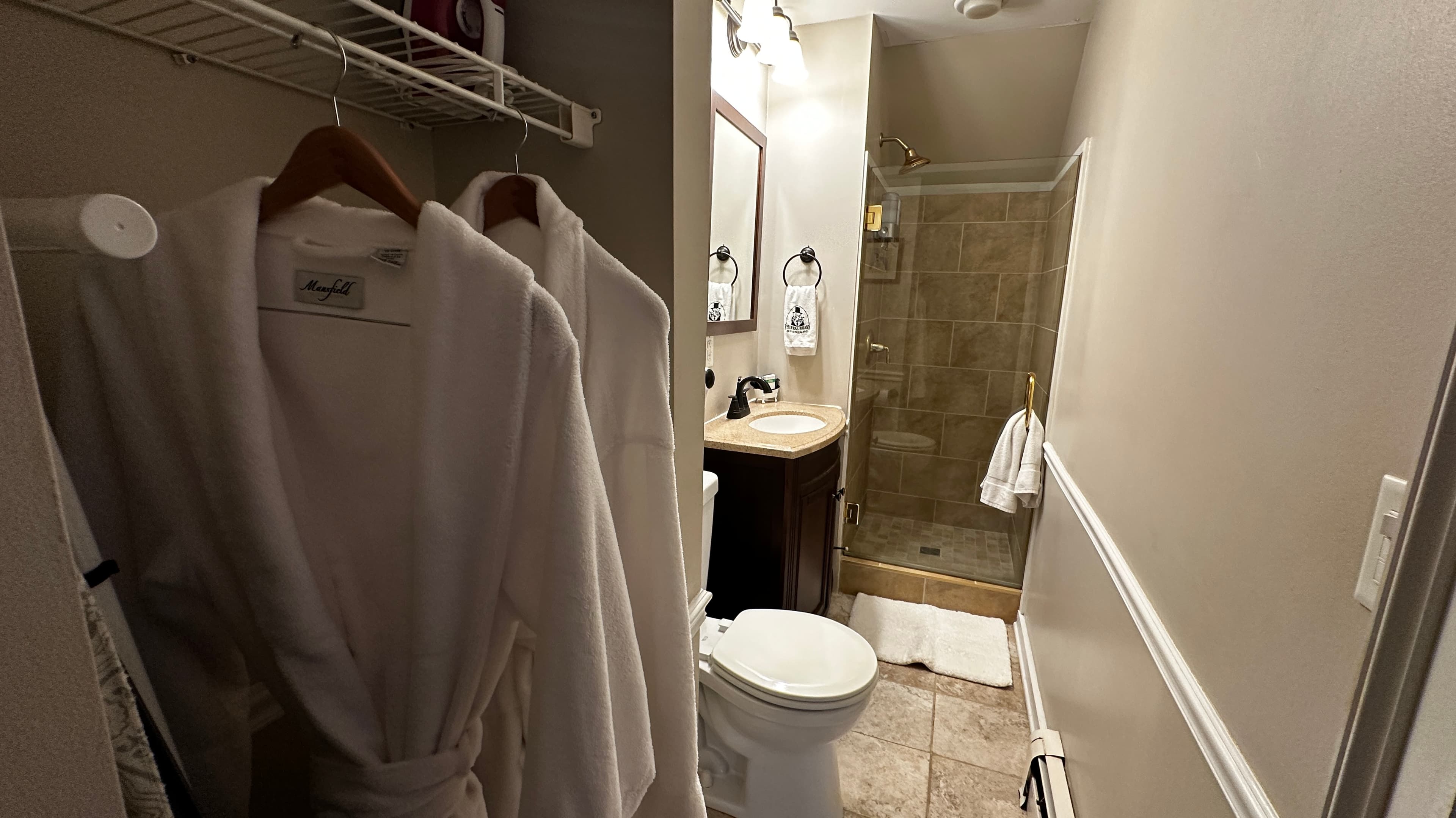 White plush bathrobes hang on a wire shelf in the foreground of a narrow bathroom. The background features a white toilet, a dark wood vanity with a white sink, and a walk-in shower with tan tiles and a glass door.