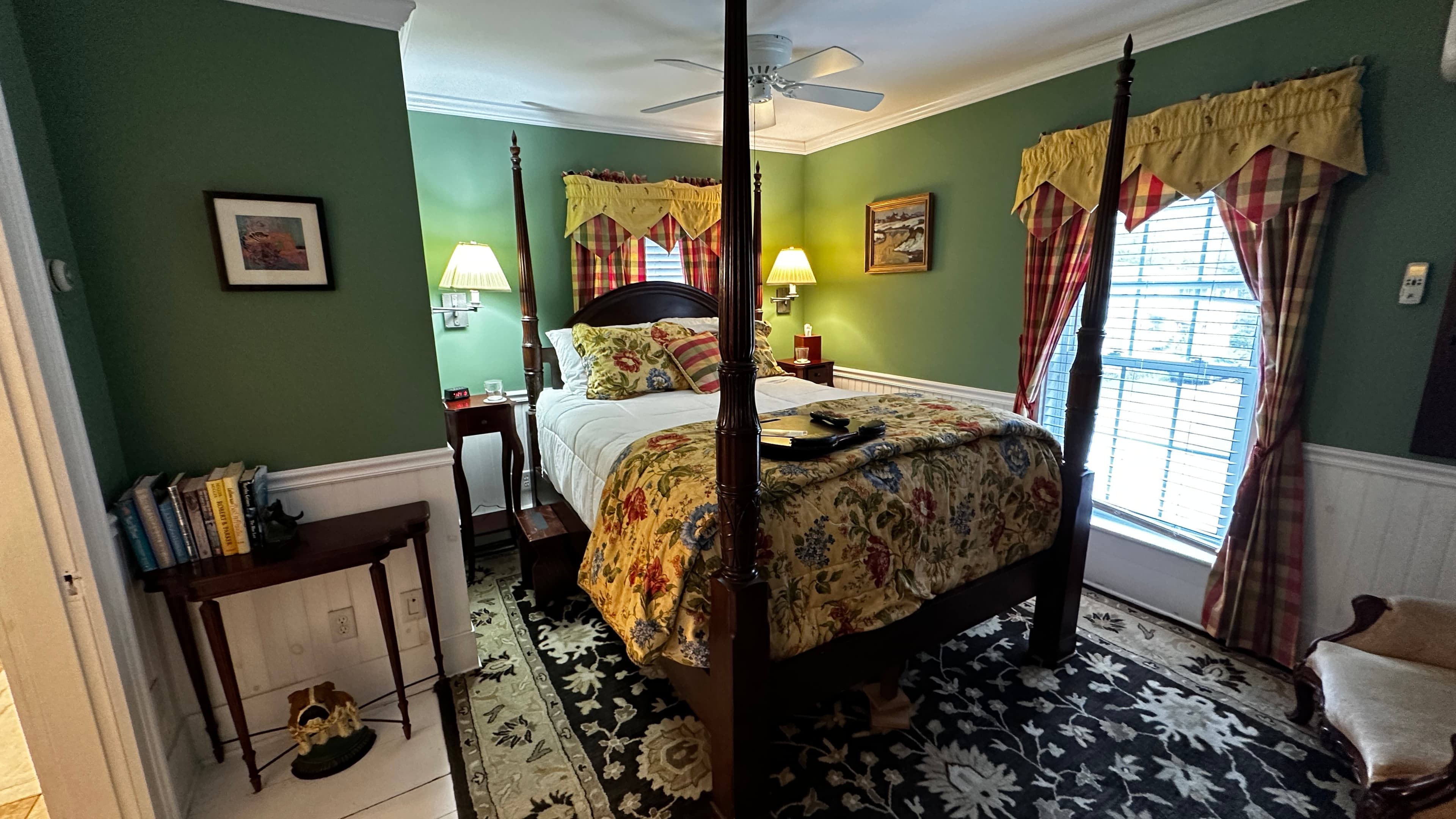 The traditional Green Room features fern-green walls, white wainscoting, and a dark wood four-poster bed with a yellow floral coverlet. The room is decorated with a patterned area rug, an antique dresser with an ornate mirror, and classic window valances.