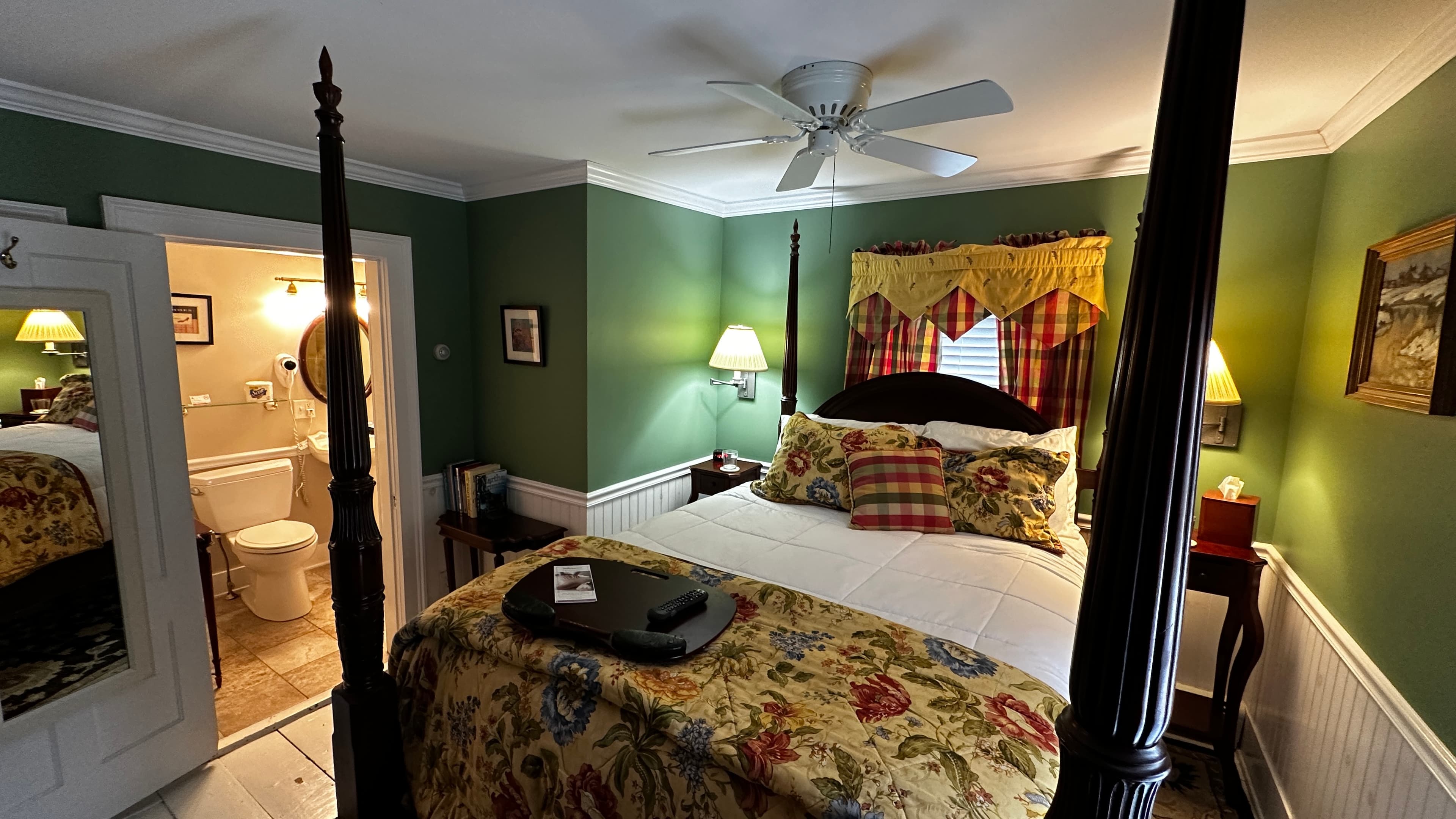The Green Room featuring fern-green walls, a dark wood four-poster bed with a yellow floral coverlet, and classic red-and-yellow plaid curtains. The room includes an antique dresser with an ornate mirror, a cream-colored armchair, and a dark patterned rug over white floors.