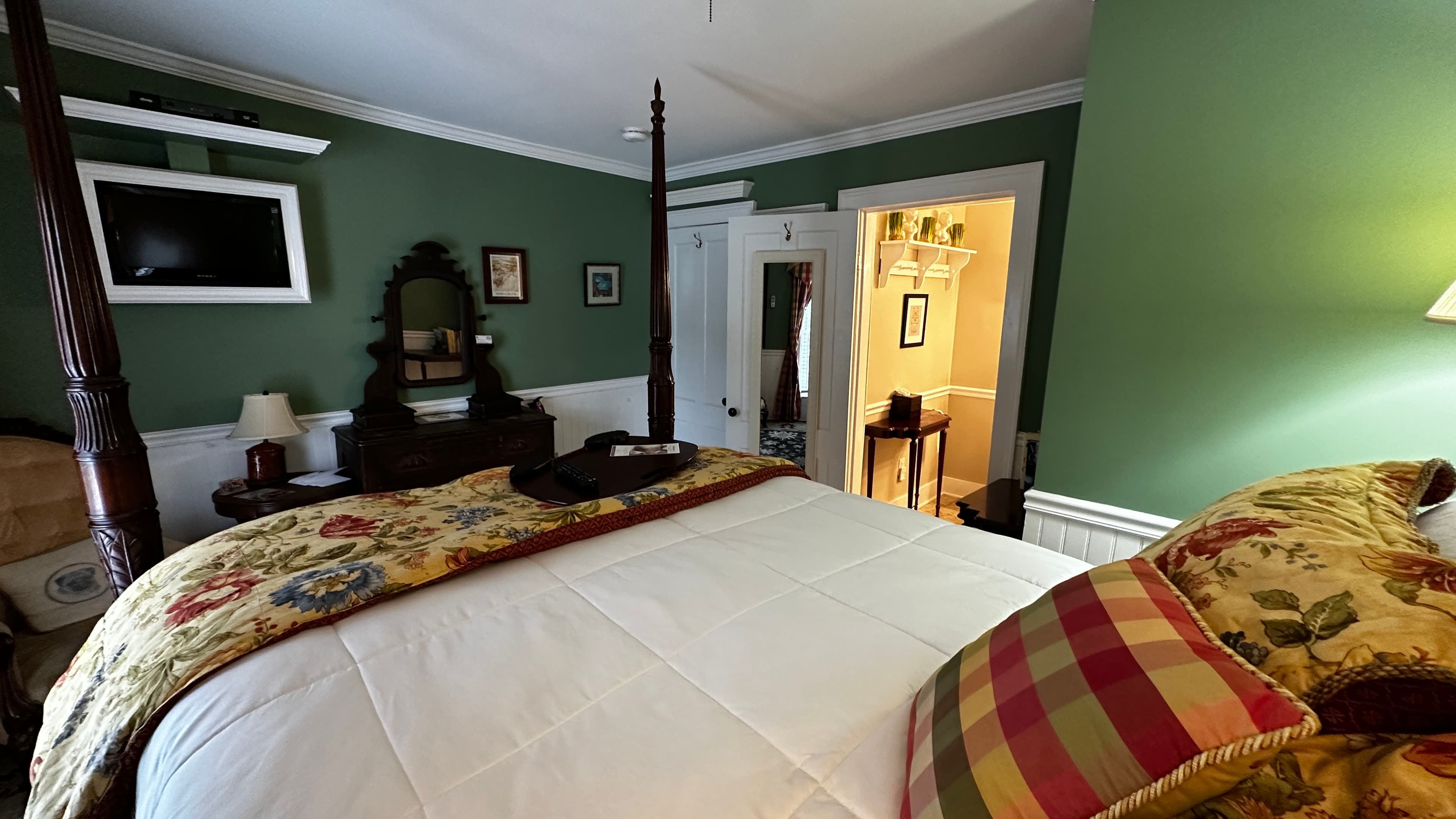 A guest room with fern-green walls, a dark wood four-poster bed with a yellow floral coverlet, and classic red-and-yellow plaid curtains. The room includes an antique dresser with an ornate mirror, a cream-colored armchair, and a dark patterned rug over white floors.