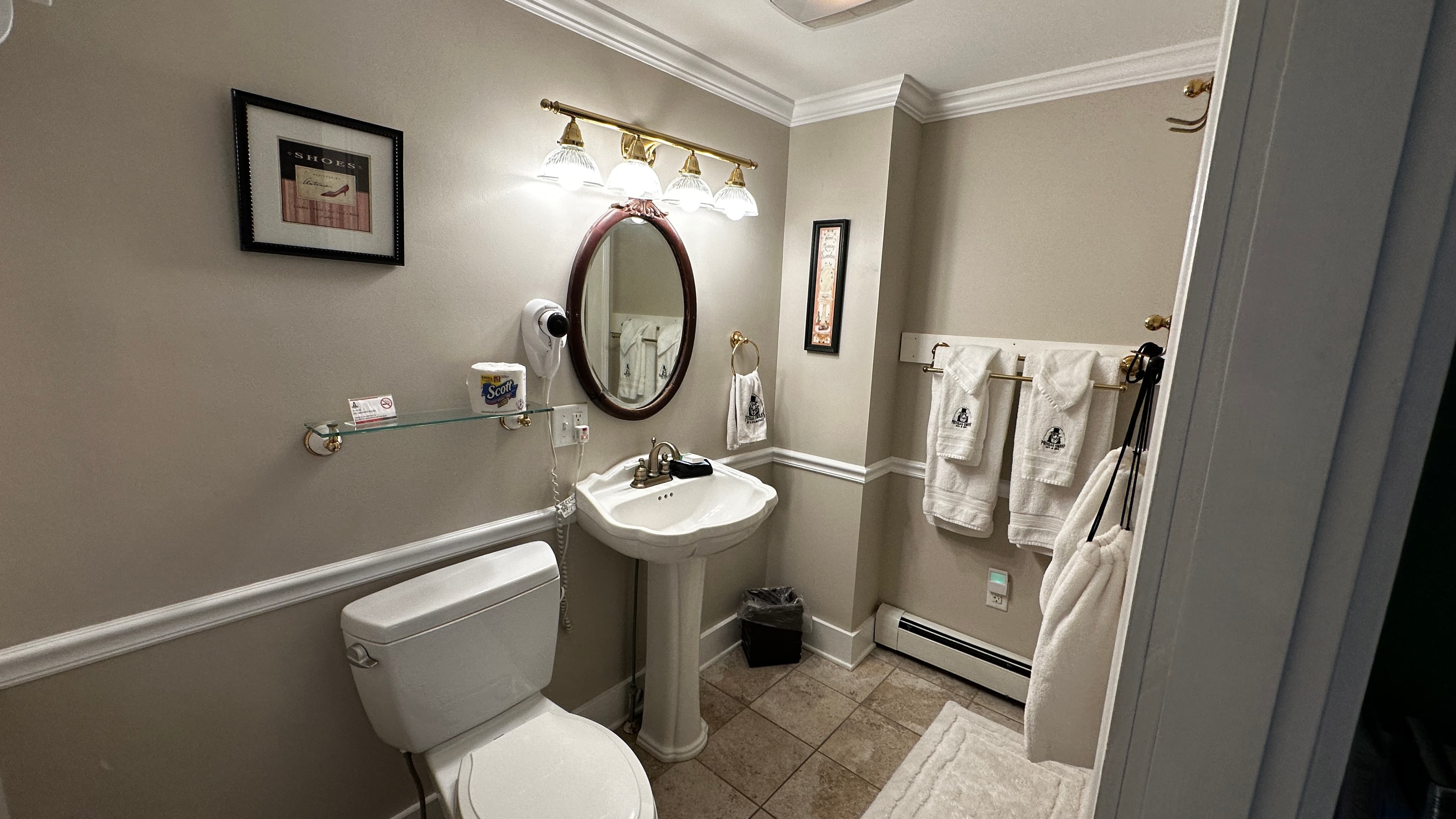 A traditional bathroom featuring a white pedestal sink with an oval mirror, a white toilet, and tan floor tiles. The walls are painted a neutral taupe with white wainscoting, and several white towels hang on a rack to the right.