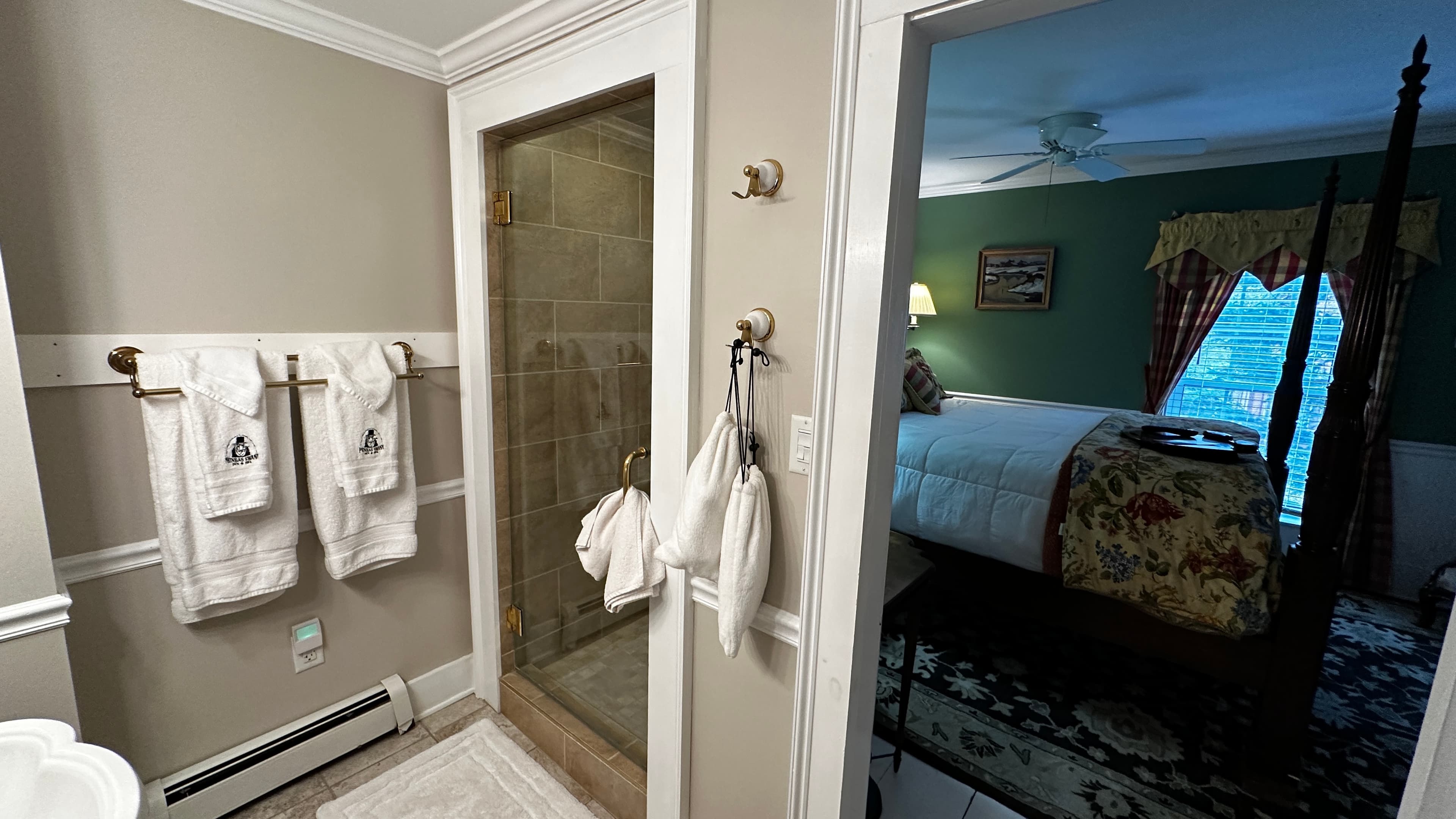 A view from the private bathroom looking back into the Green Room, showing white towels branded with the Phineas Swann logo hanging on the wall. The doorway reveals the dark wood four-poster bed with its yellow floral coverlet and the room's distinctive fern-green walls.
