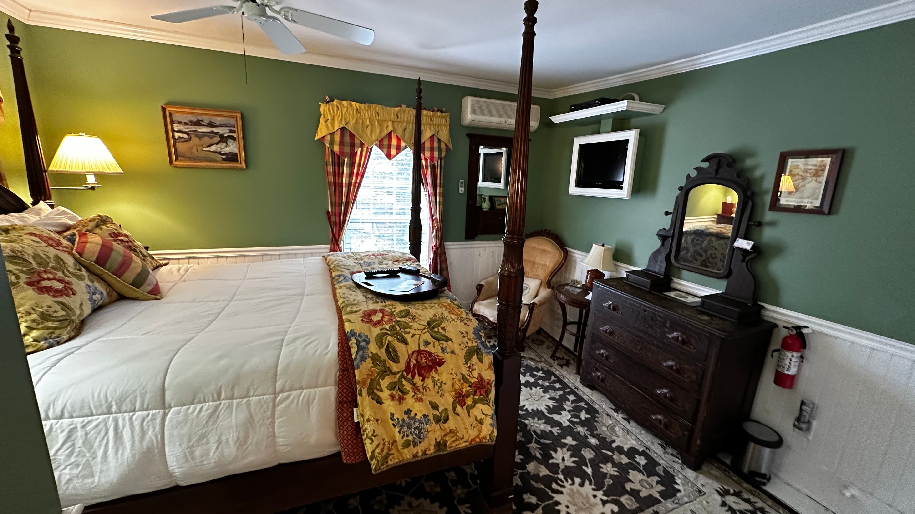 The Green Room featuring a dark wood four-poster bed with a vibrant yellow and red floral coverlet. The room features fern-green walls, white beaded wainscoting, an antique dresser with a mirror, and a classic wingback chair.