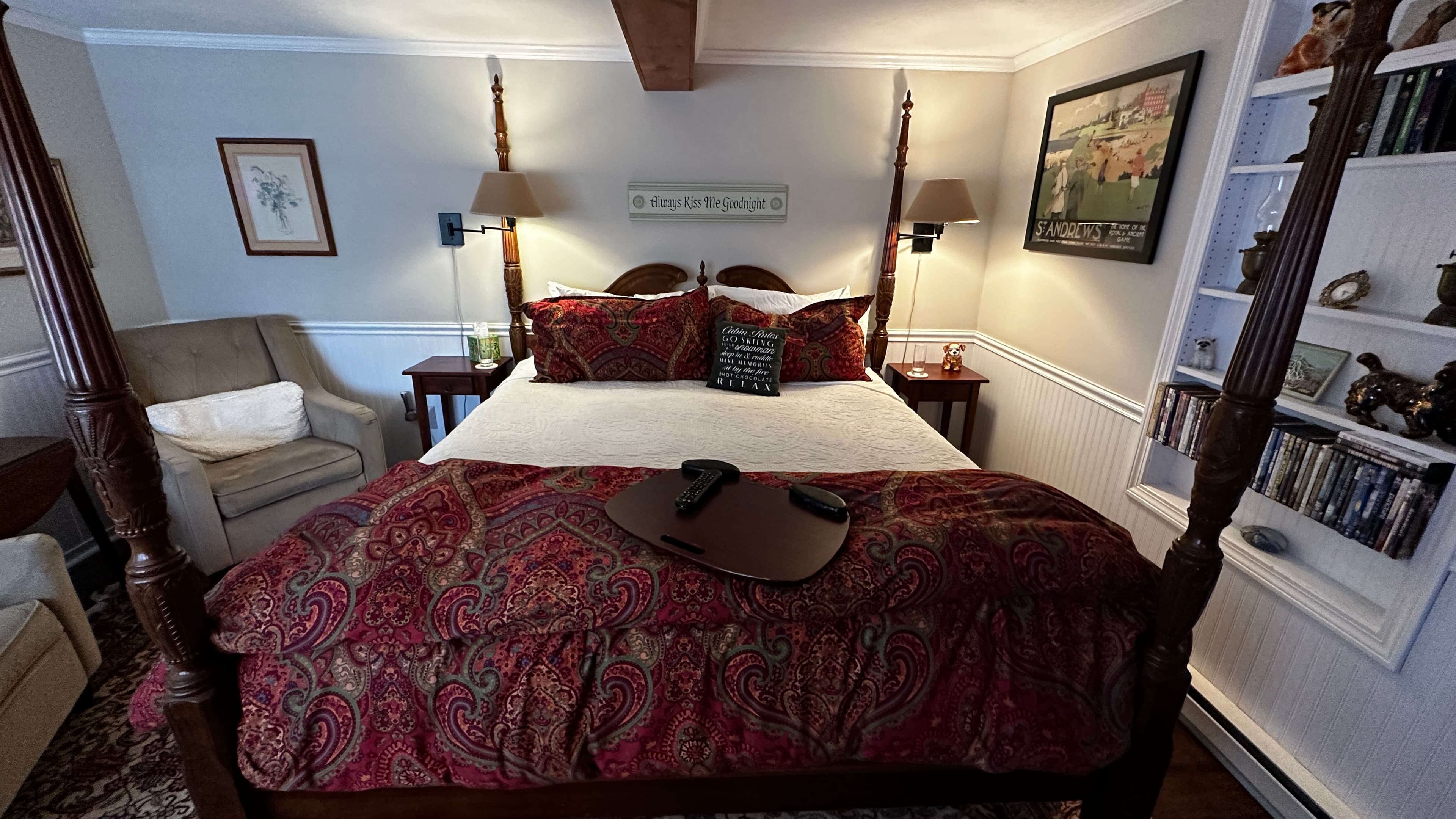 A guest bedroom featuring a four-poster king-size bed with white linens and a red patterned throw. The room includes two plush gray reading chairs, a decorative patterned area rug over wood floors, a wall-mounted TV, and built-in bookshelves.