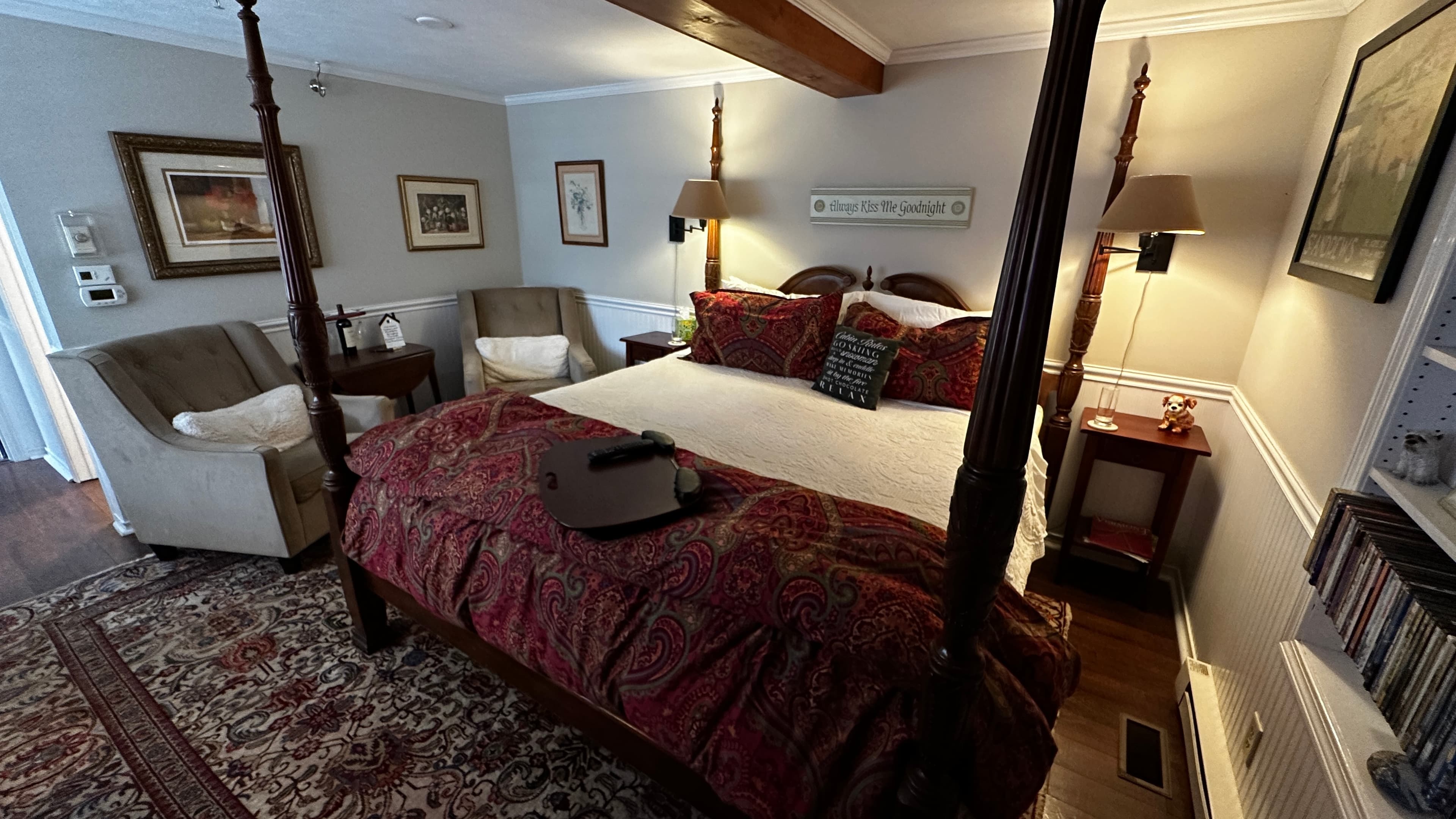 A cozy guest bedroom featuring a dark wood four-poster king-size bed with crisp white linens and a decorative red paisley throw. The room includes two plush gray armchairs, a traditional patterned area rug over dark wood floors, and warm lighting from bedside lamps.