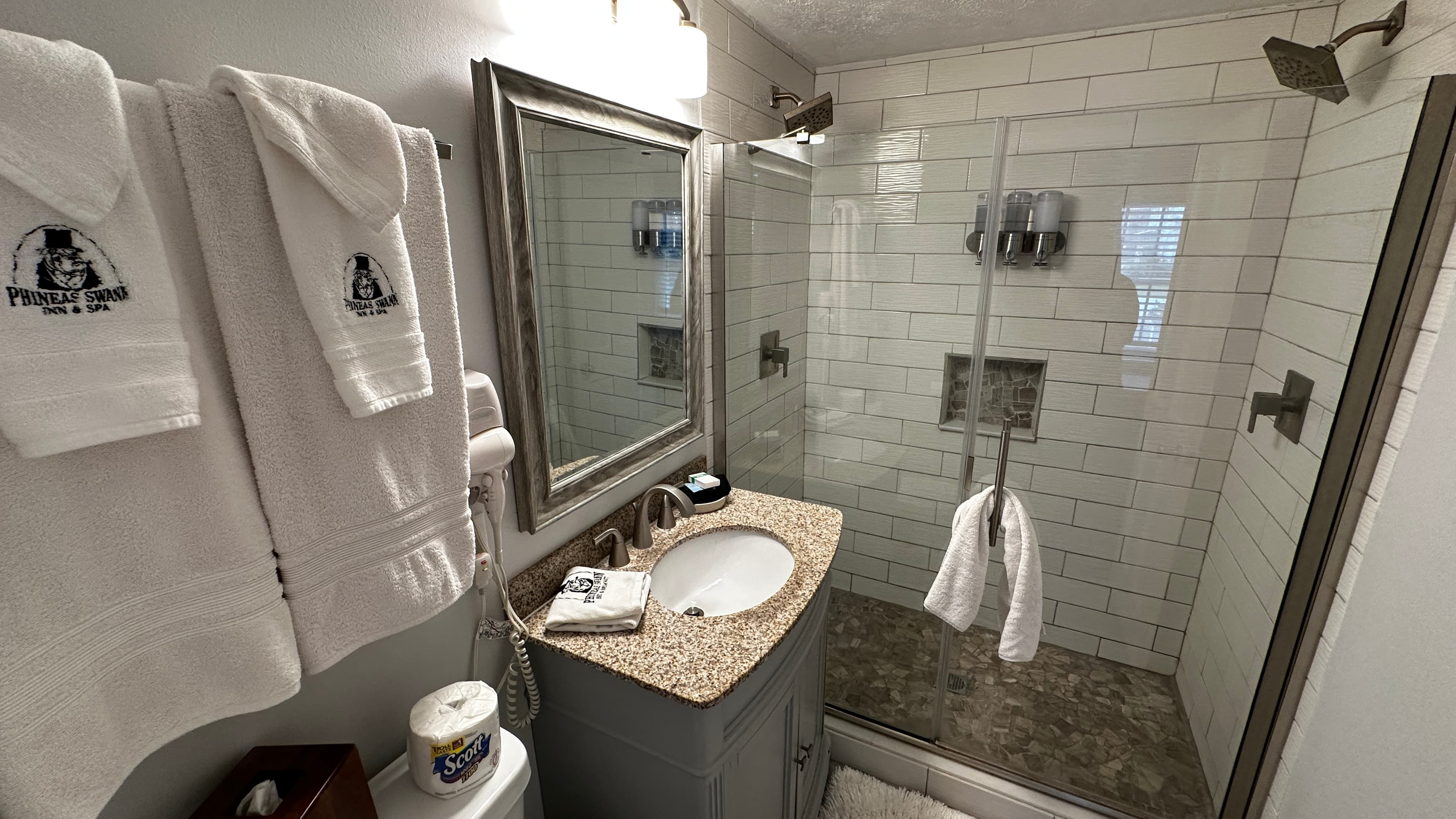 A modern bathroom features a white subway-tiled walk-in shower with a glass door and stone-tiled floor. Next to the shower is a grey vanity with a speckled granite countertop, a white oval sink, and an ornate silver-framed mirror.