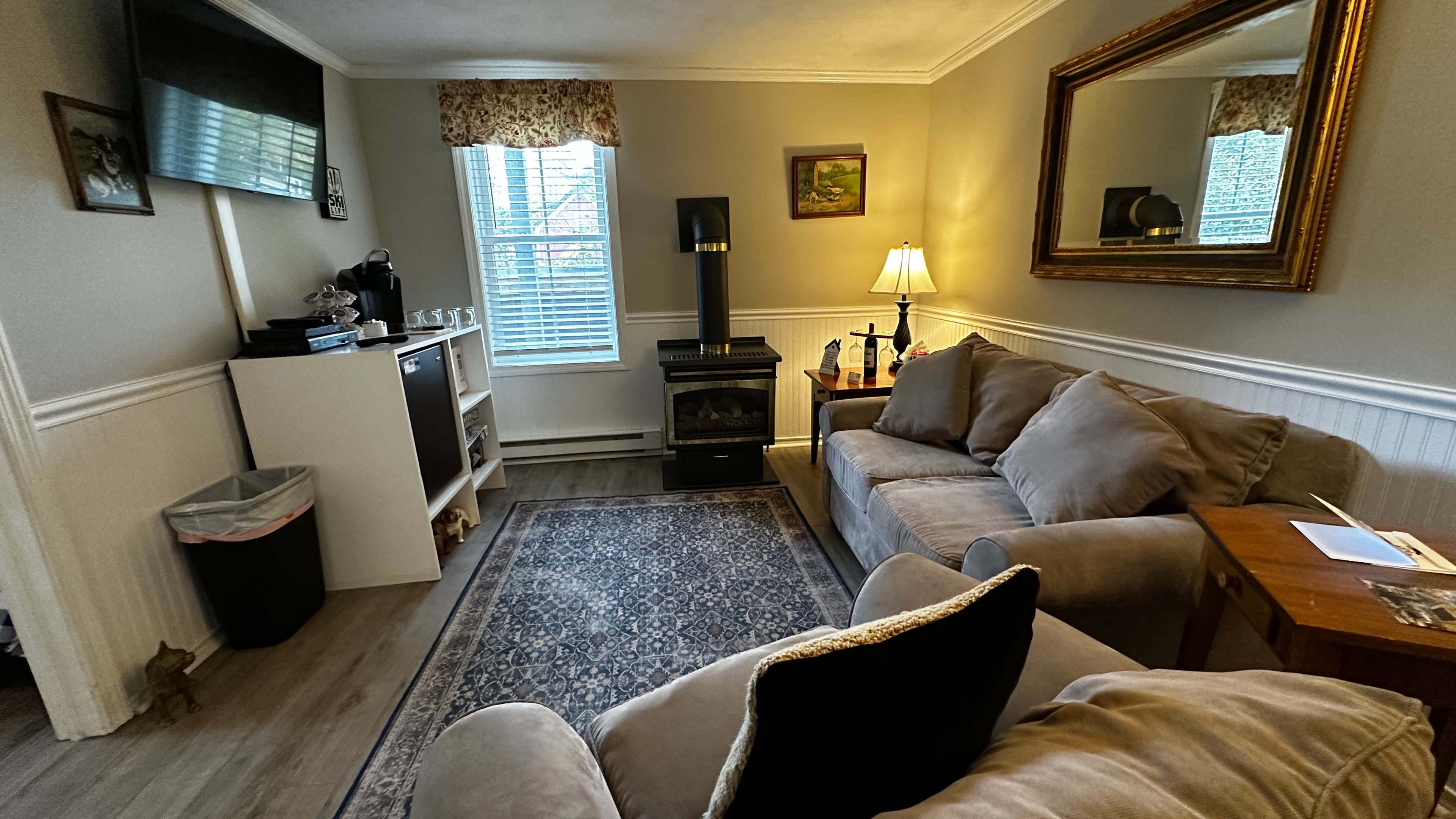 A comfortable living area in a suite featuring a grey sofa with plush pillows, a patterned area rug, and a small kitchenette station with a mini-fridge and coffee maker. The room is decorated with a large gold-framed mirror, a black gas log fireplace, and wainscoting along the lower half of the walls.